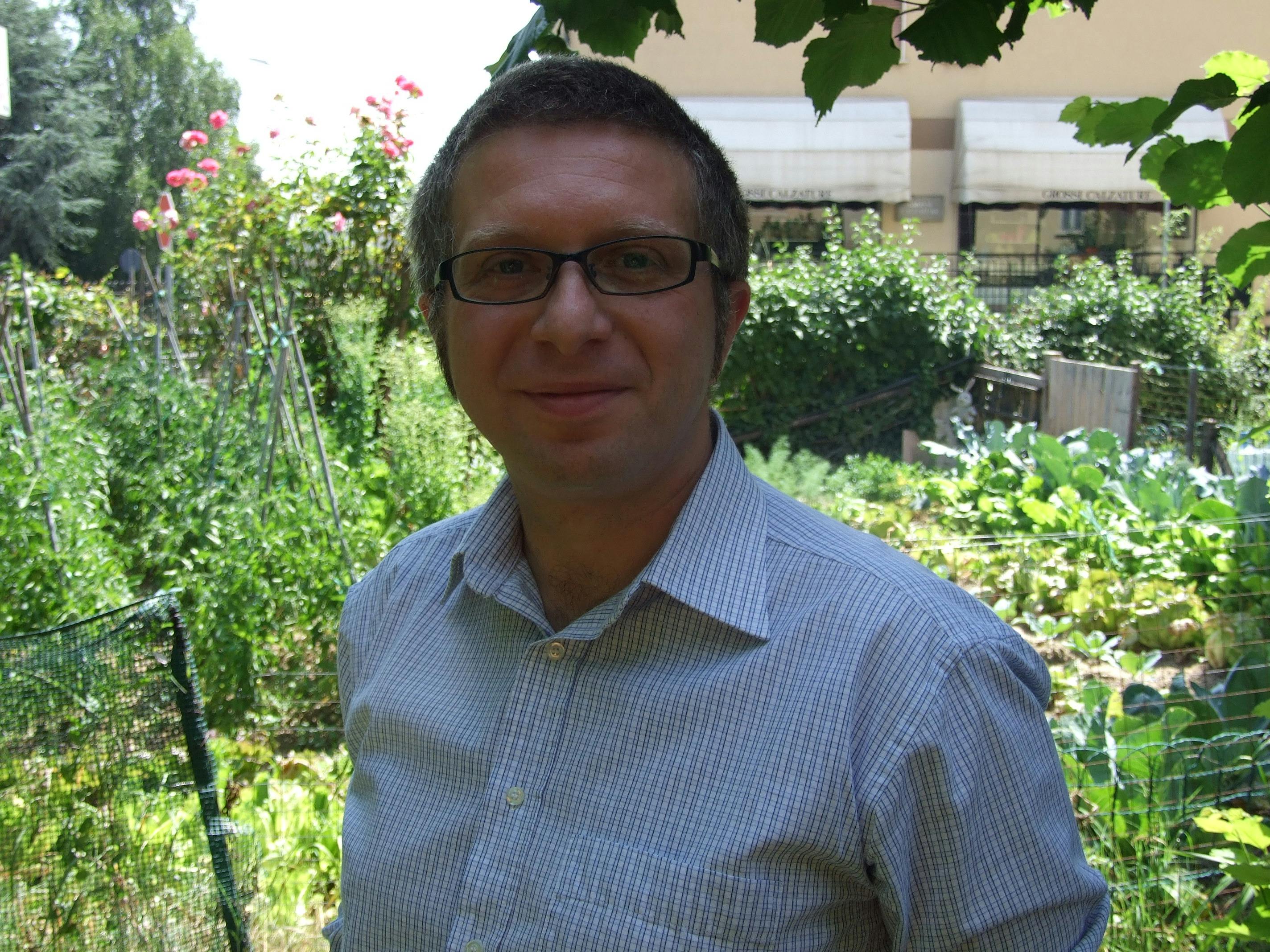 Ritratto di Massimo De Maio. Indossa una camicia azzurra e degli occhiali. Sullo sfondo un orto verde.