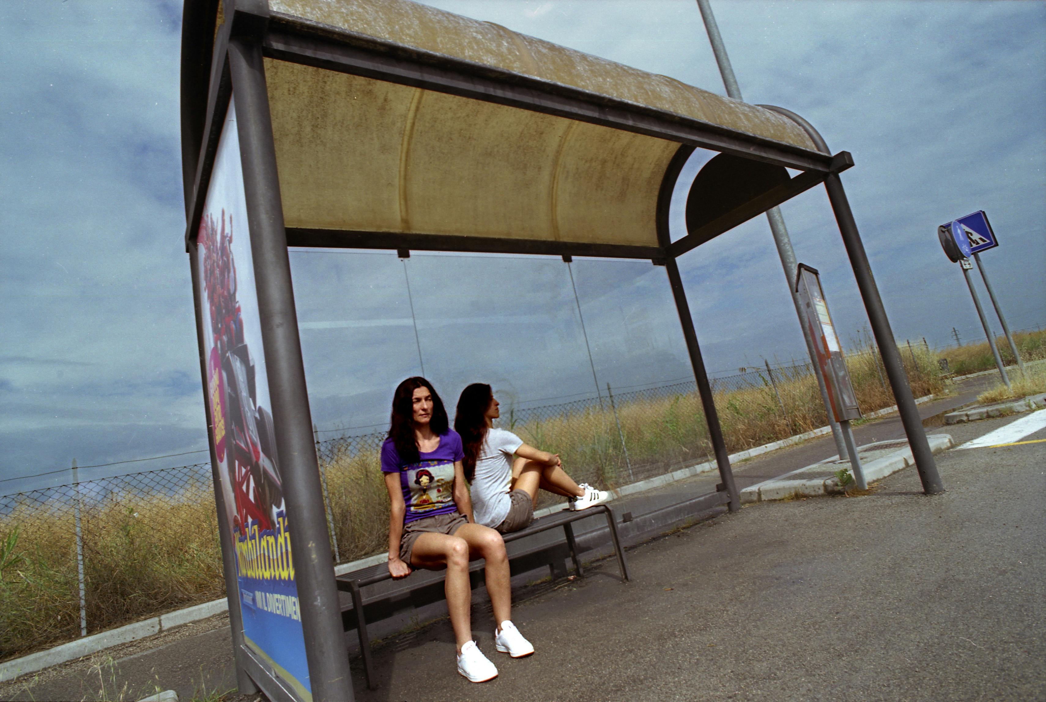 Two artists are sitting at the bus stop. The sky is cloudy. One is looking straight ahead, while the other is sitting at a 90° angle to the first.