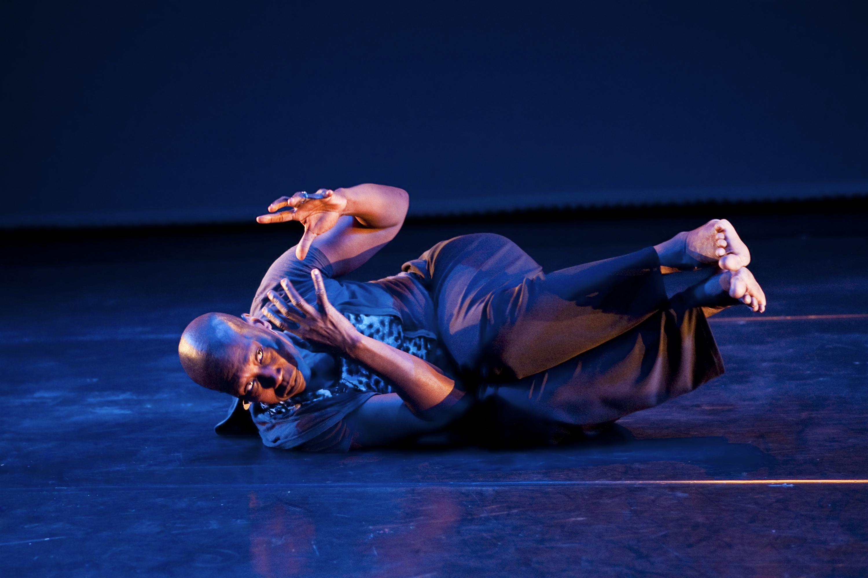 A dancer is lying on the ground, on their side. They hold their knees close to their chest, with their feet suspended in the air.