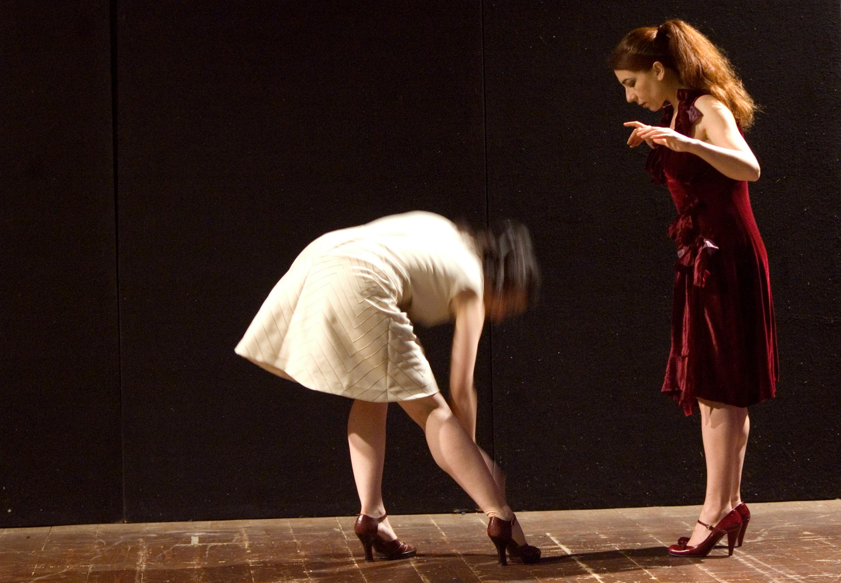 Due ballerine, entrambe con i tacchi. La ballerina a sinistra è vestita di bianco e si piega in avanti. Quella a destra è vestita di rosso e guarda la prima ballerina.