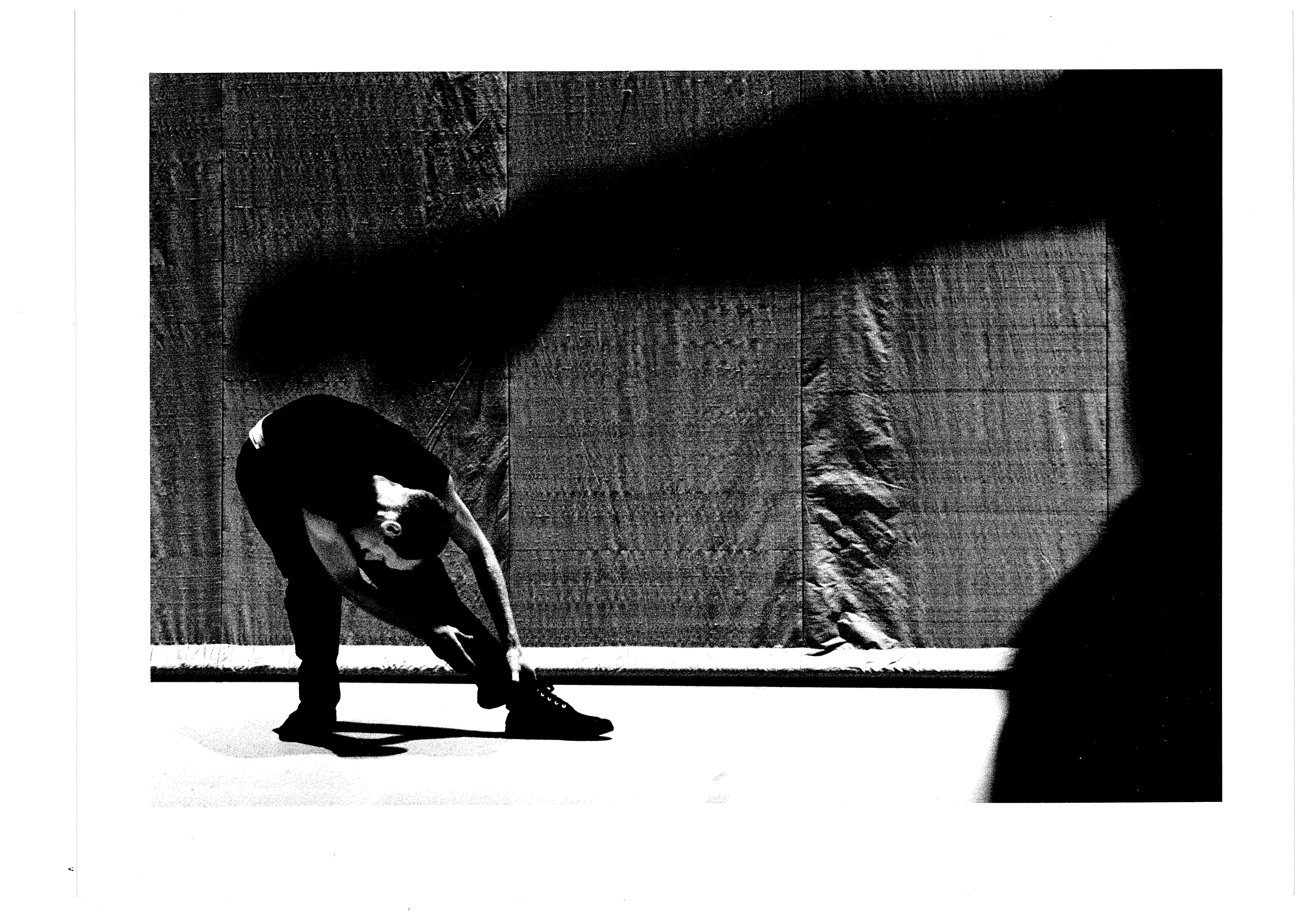 a dancer bent towards the ankles, with a blurred close-up of an arm in the foreground