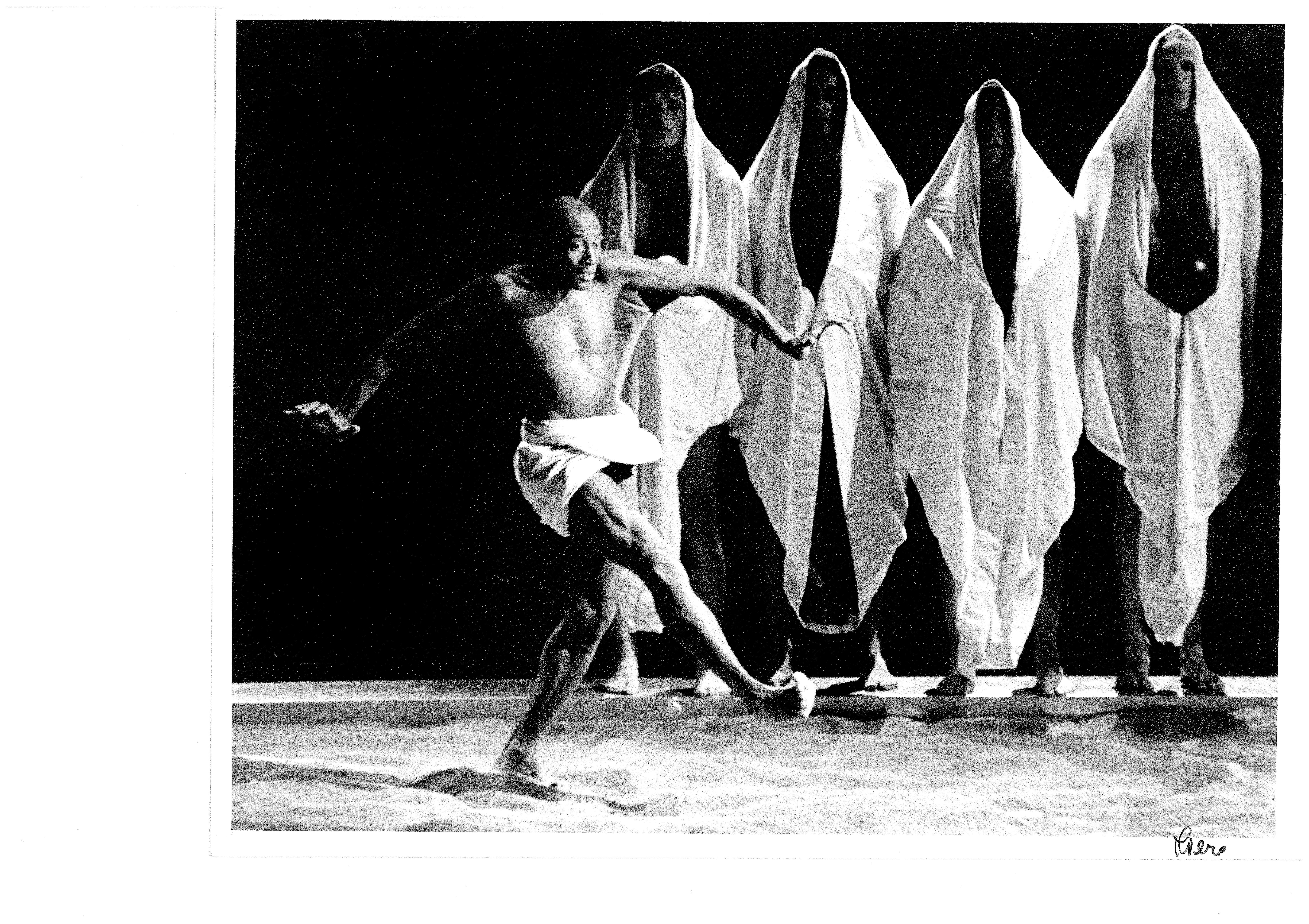 Five people. In the foreground, a shirtless man walks with his arms outstretched. Behind him, four people are standing in white tunics.