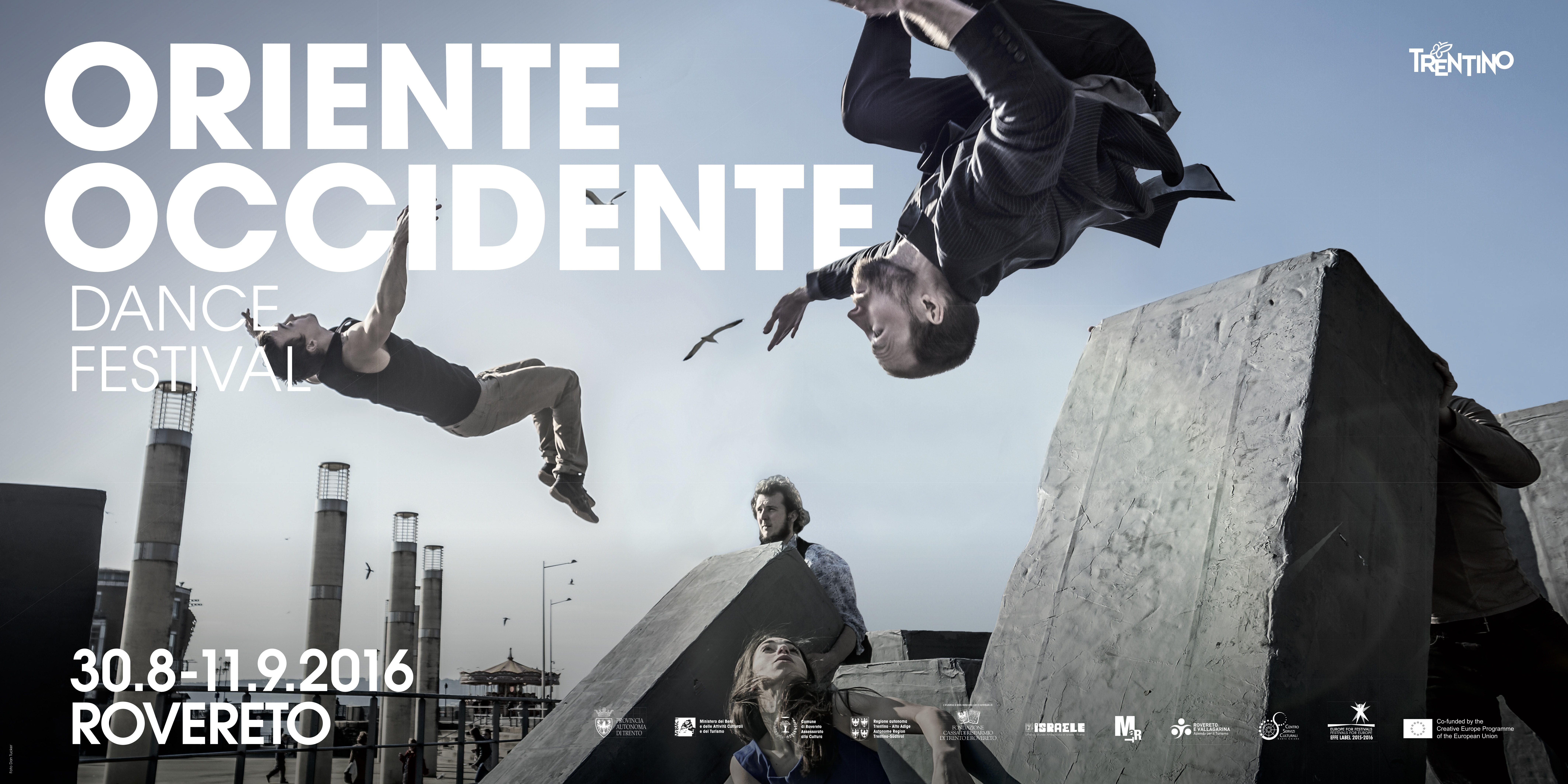 Lavoro grafico del Festival: testi bianchi (Oriente Occidente Dance Festival), sullo sfondo uno scorcio di città e blocchi di cemento, due figure fanno acrobazie e altre due sono ferme tra i blocchi.