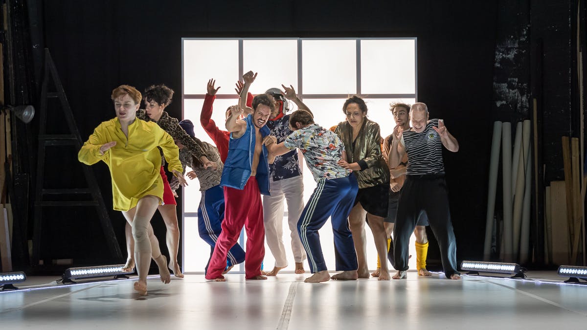 Dieci ballerini con costumi colorati catturati durante una coreografia in uno spazio chiuso con pavimento e luci bianche