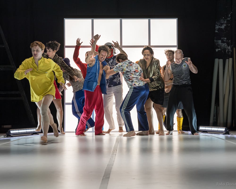 Dieci ballerini con costumi colorati catturati durante una coreografia in uno spazio chiuso con pavimento e luci bianche