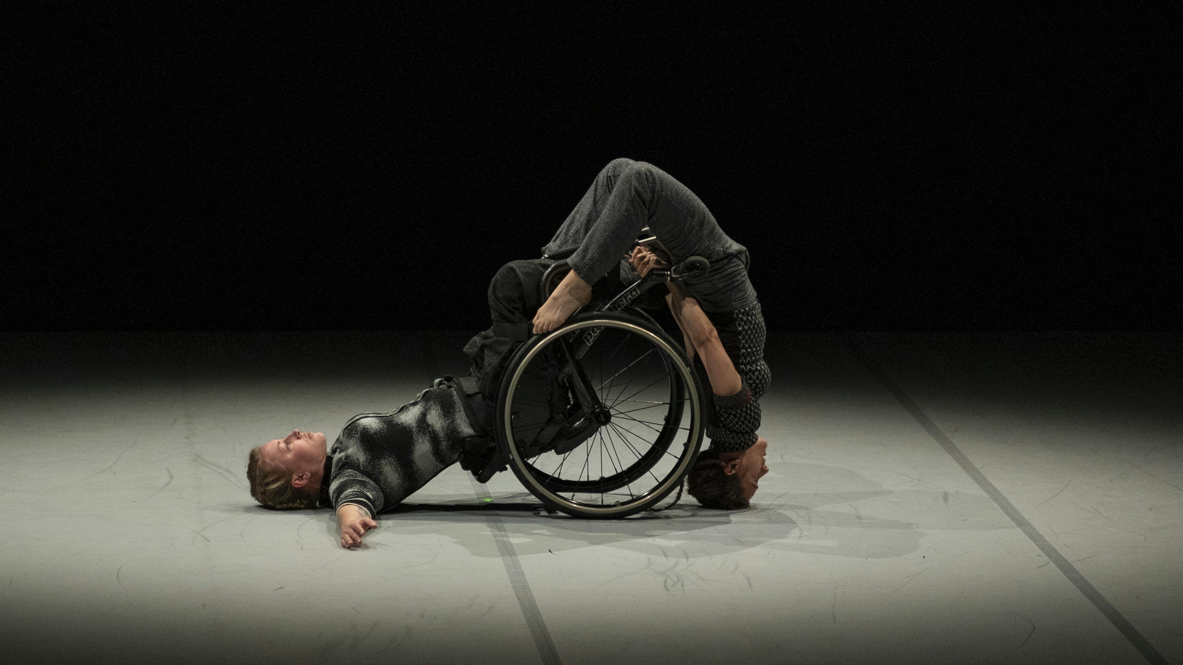 A dancer using a wheelchair leans on her back. another dancer balances on her head using the wheelchair for support
