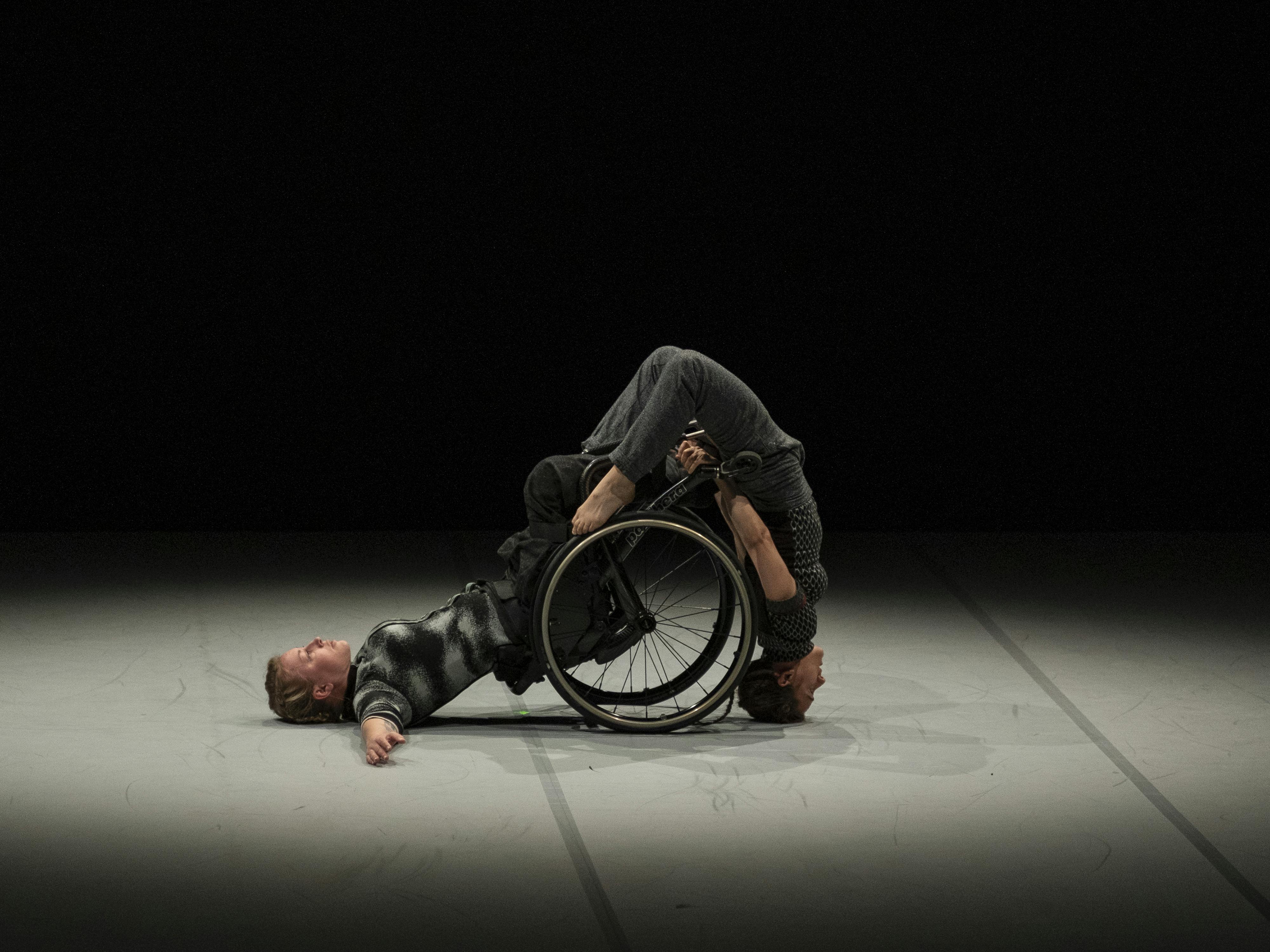 A dancer using a wheelchair leans on her back. another dancer balances on her head using the wheelchair for support
