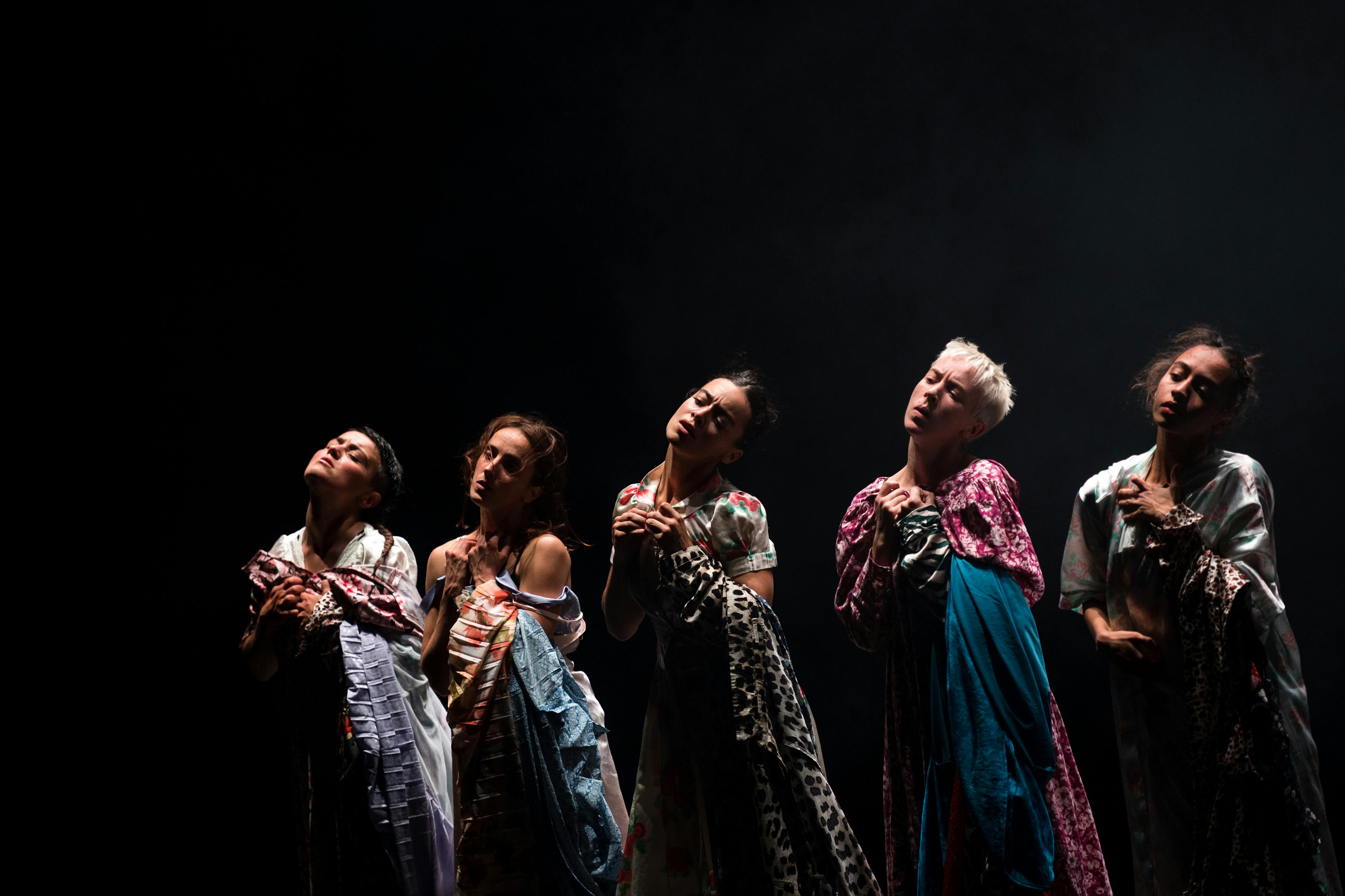 Five dancers stand with their eyes closed and heads tilted. they wear long, colourful dresses