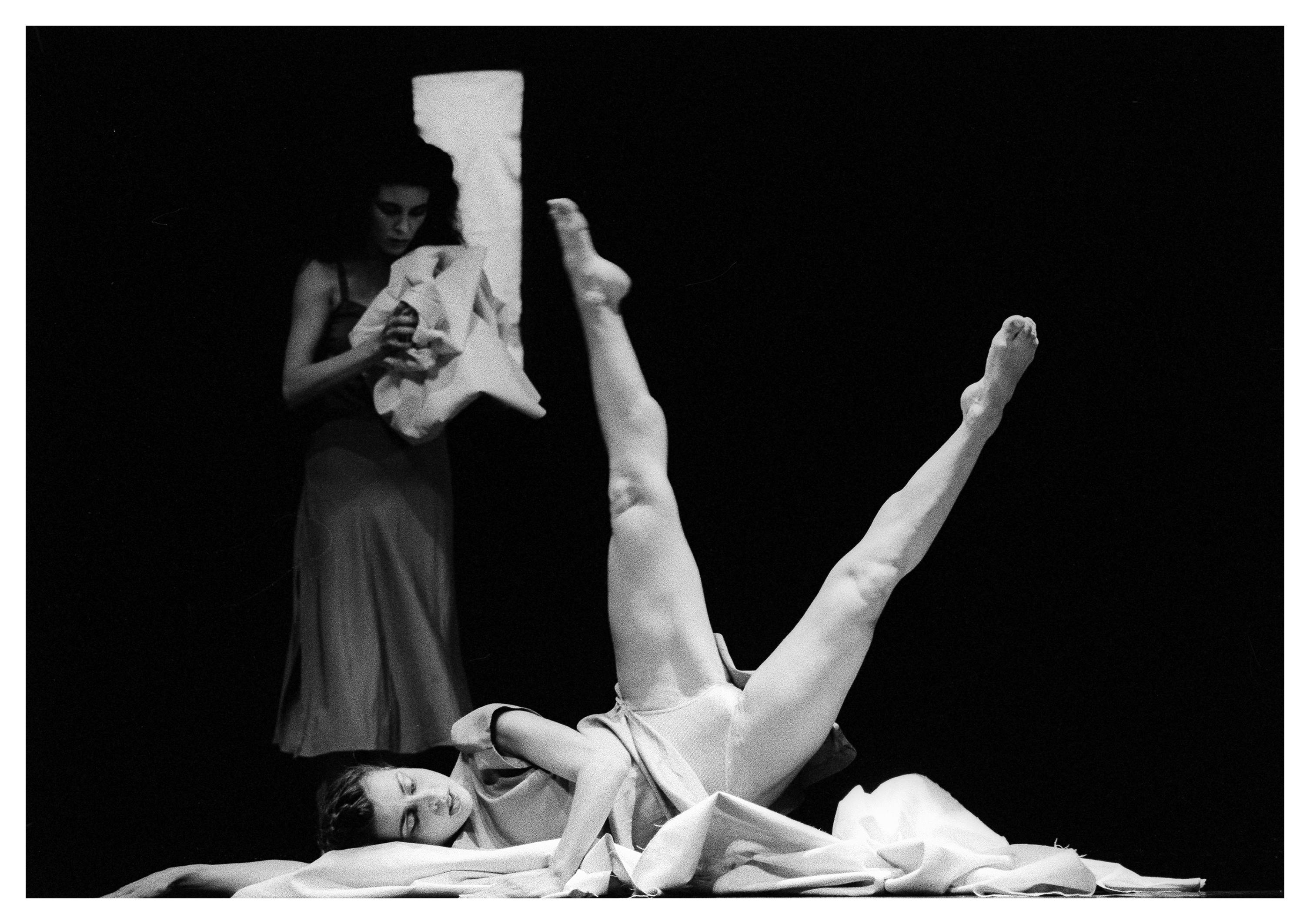 Two people on stage. One is lying on the ground with her legs stretched up, the other is standing behind her.