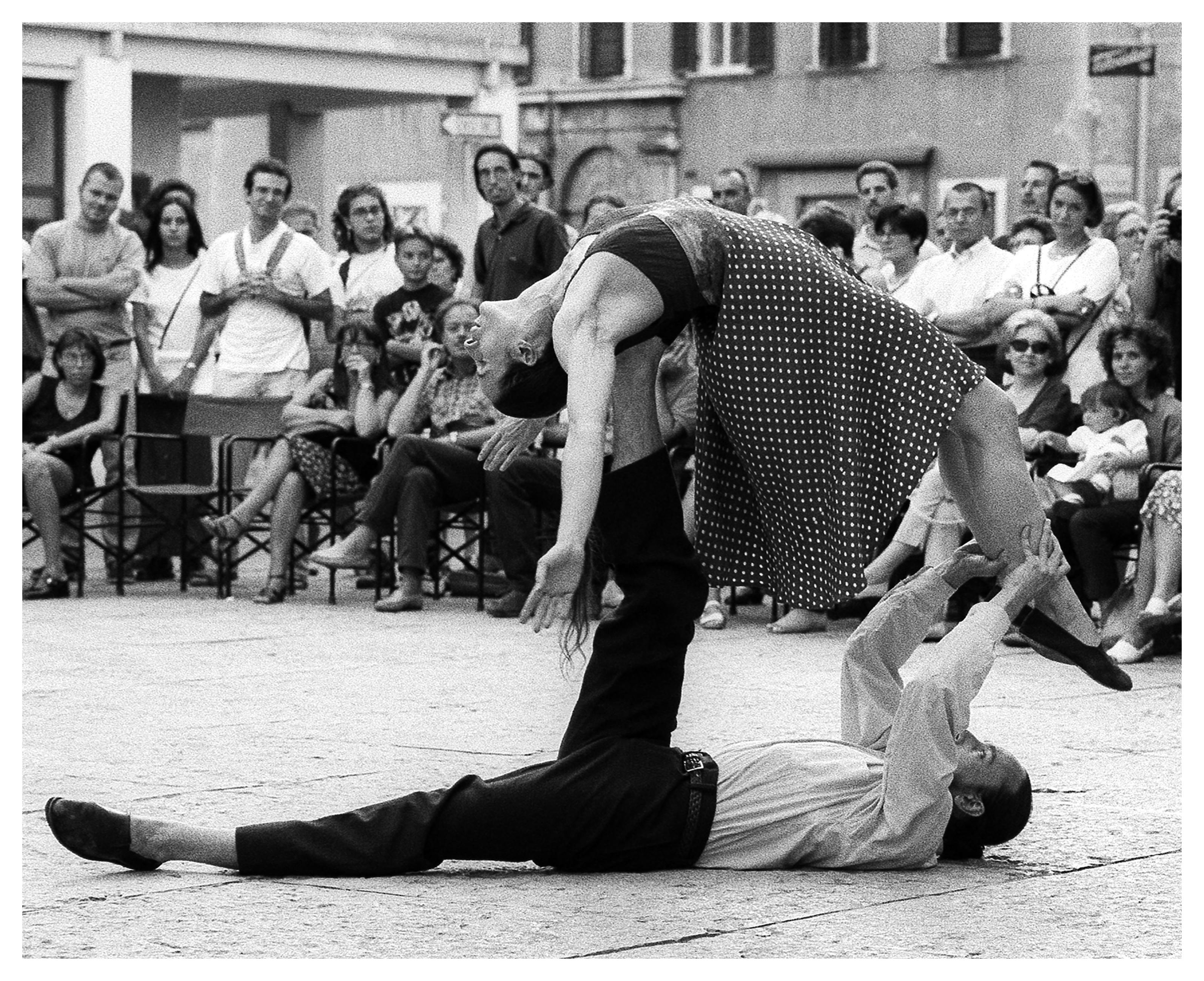 Un uomo è sdraiato in terra. Sopra di lui, una donna in equilibrio appoggiata alla gamba destra di lui. Sullo sfondo una folla che guarda.