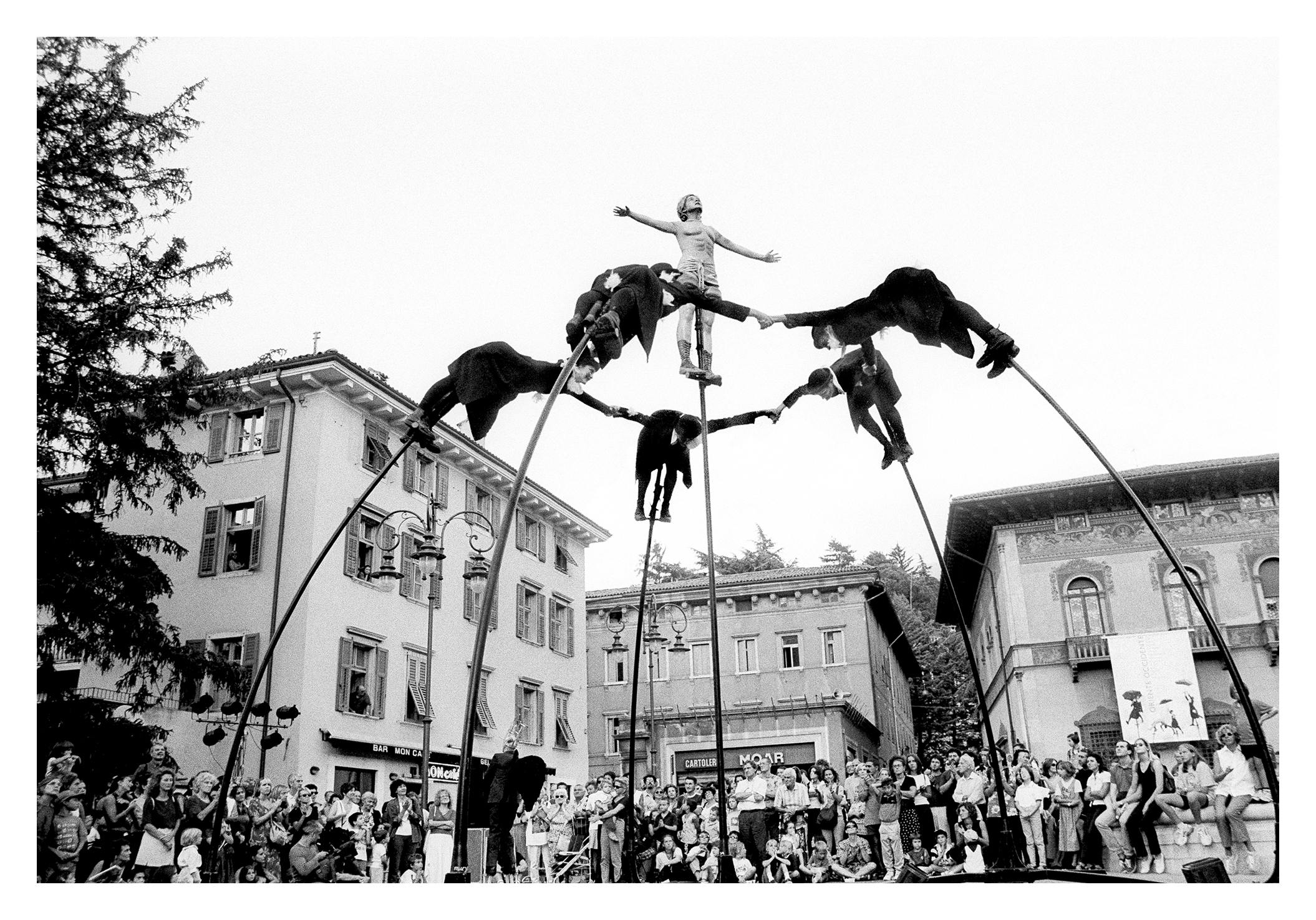 Six performers on stilts in the middle of a crowd of people. Five of them stand in a circle and hold each other for less, in the middle of the circle the sixth performer.