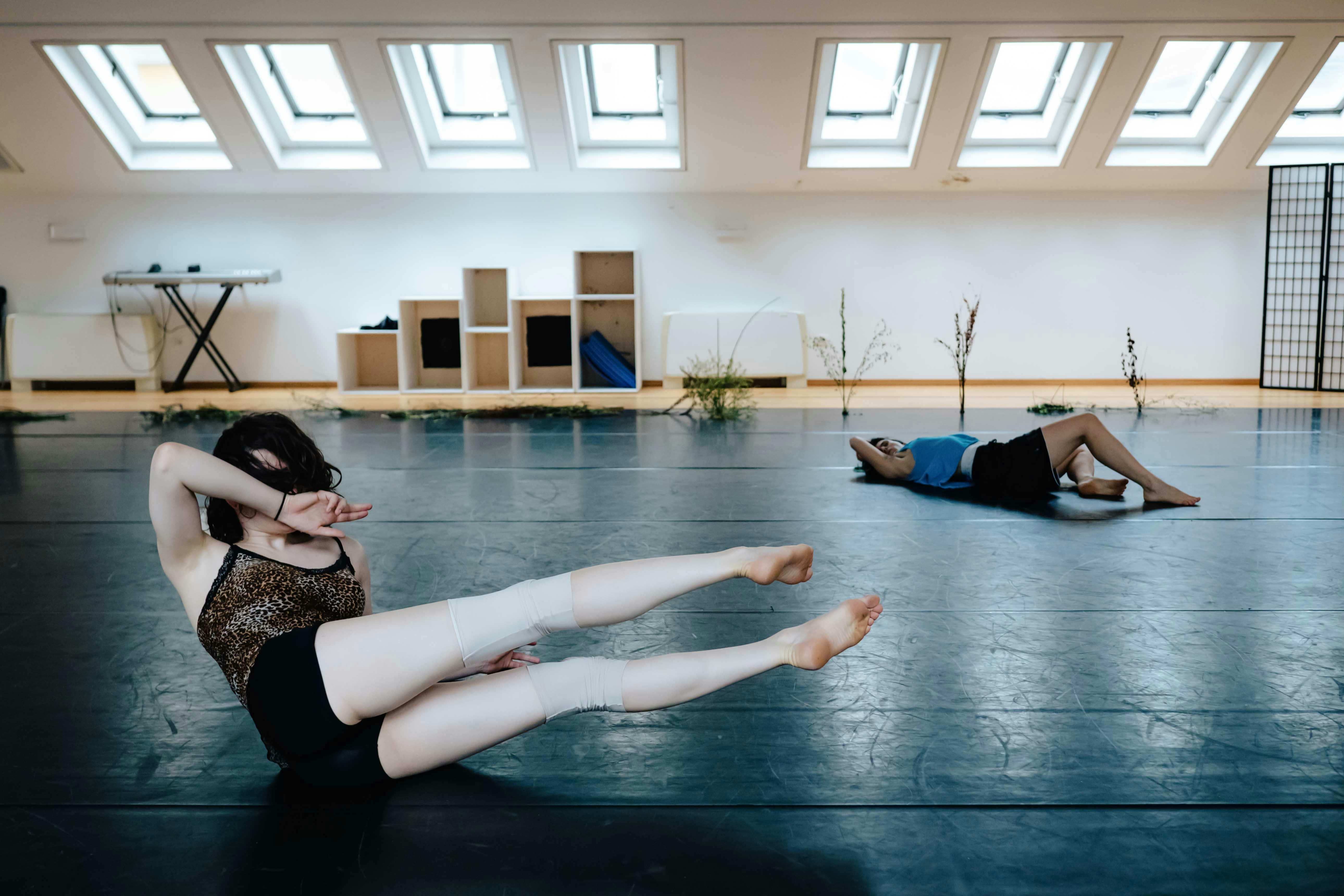 Due performer a terra, una in primo piano alza le gambe e il busto, la seconda sullo sfondo piega le gambe