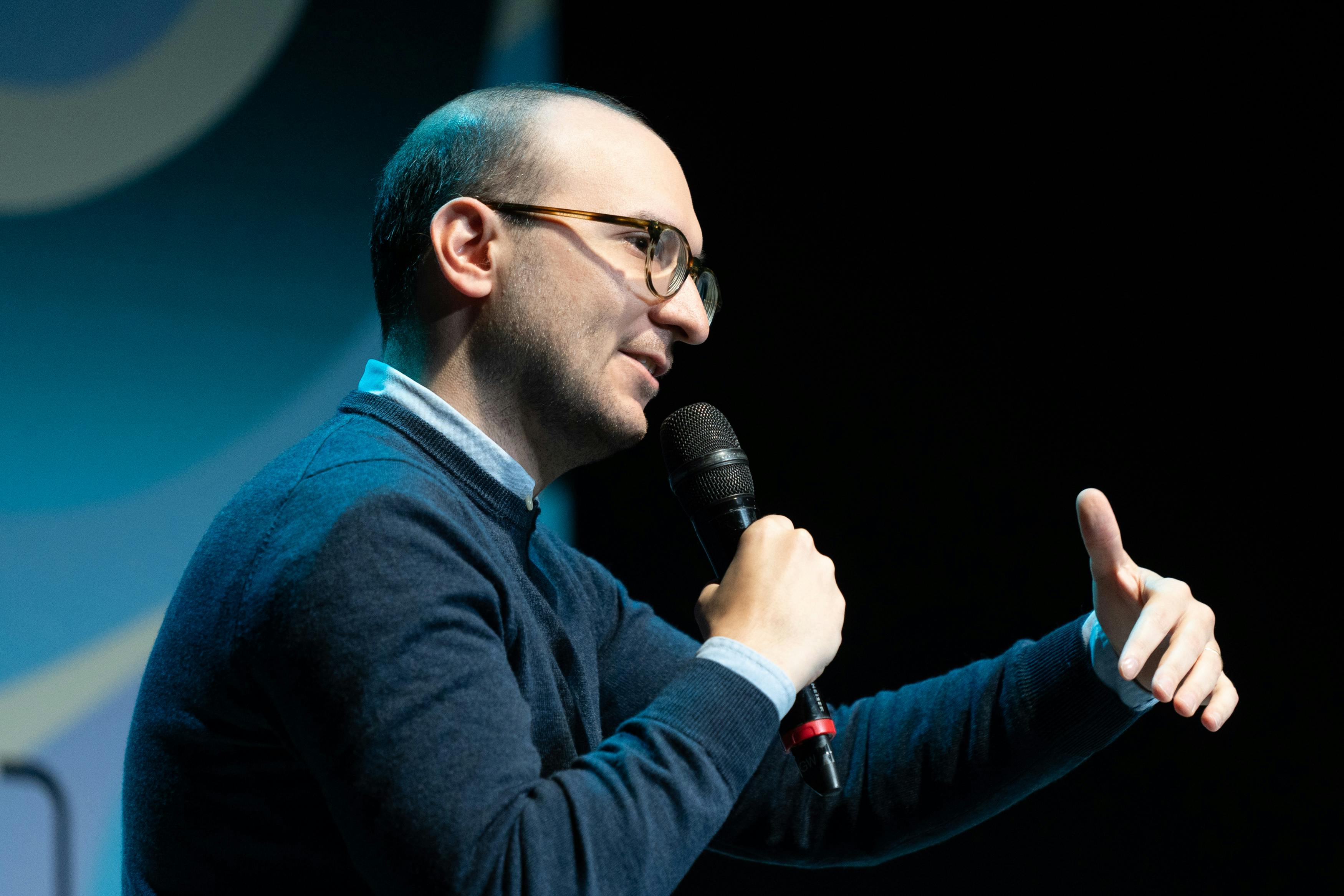 Il giornalista Luca Misculin parla con un microfono in mano. Indossa un maglione blu, camicia azzurra e un paio di occhiali da vista.
