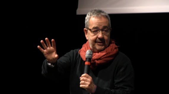 Il relatore Raffale Crocco parla mentre ha nella mano sinistra un microfono e la mano destra davanti a sè. Indossa un maglione nero e una sciarpa rossa. Ha capelli cori grigi e porta gli occhiali.