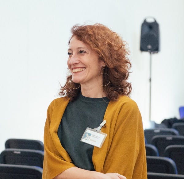 Foto della relatrice Anna Chiara Cimoli. Guarda verso la propria destra sorridendo. Indossa una maglietta grigia e un cardigan color ocra. Ha capelli fino alle spalle rossi.
