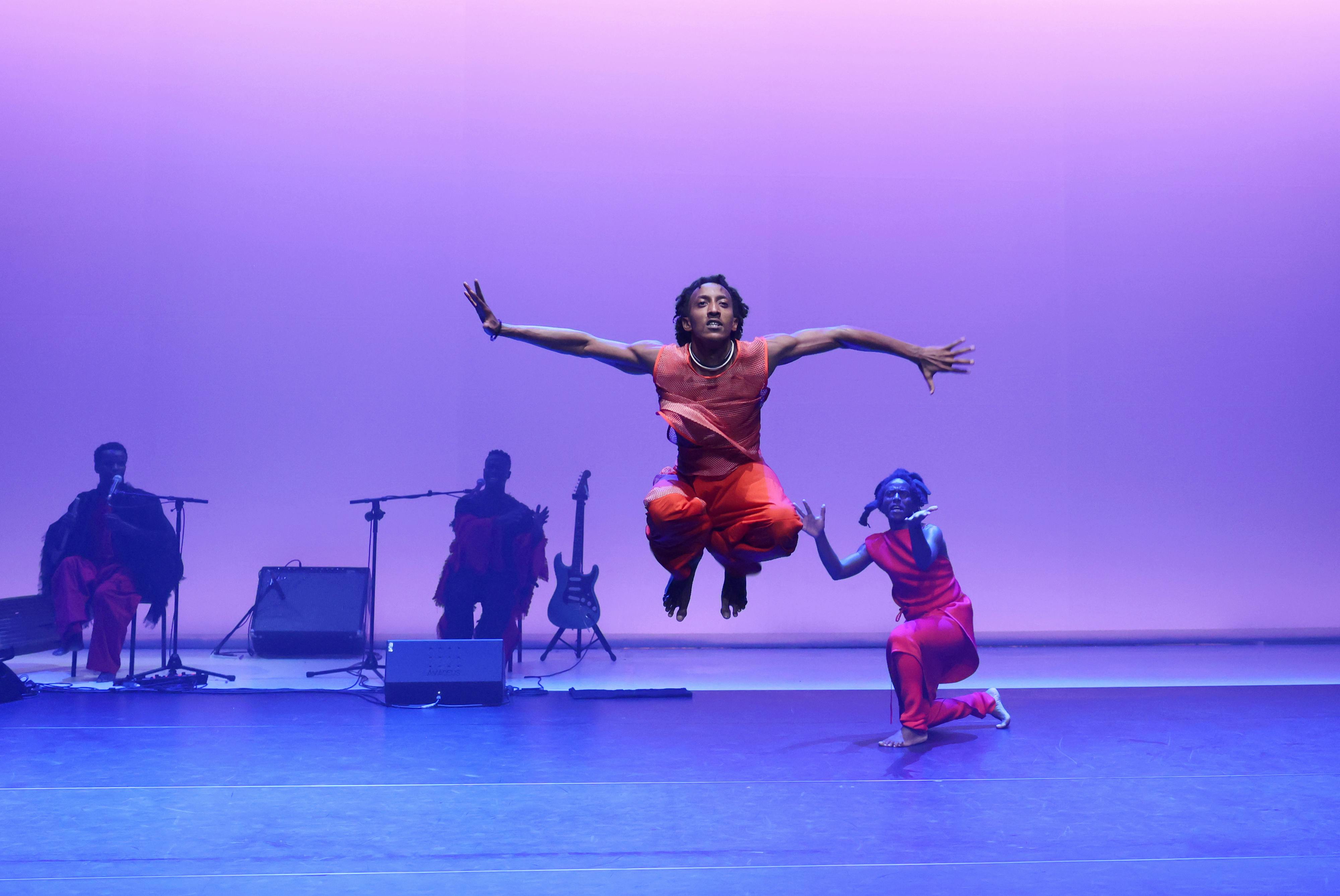 Un danzatore salta su un palco illuminato di viola davanti a dei musicisti