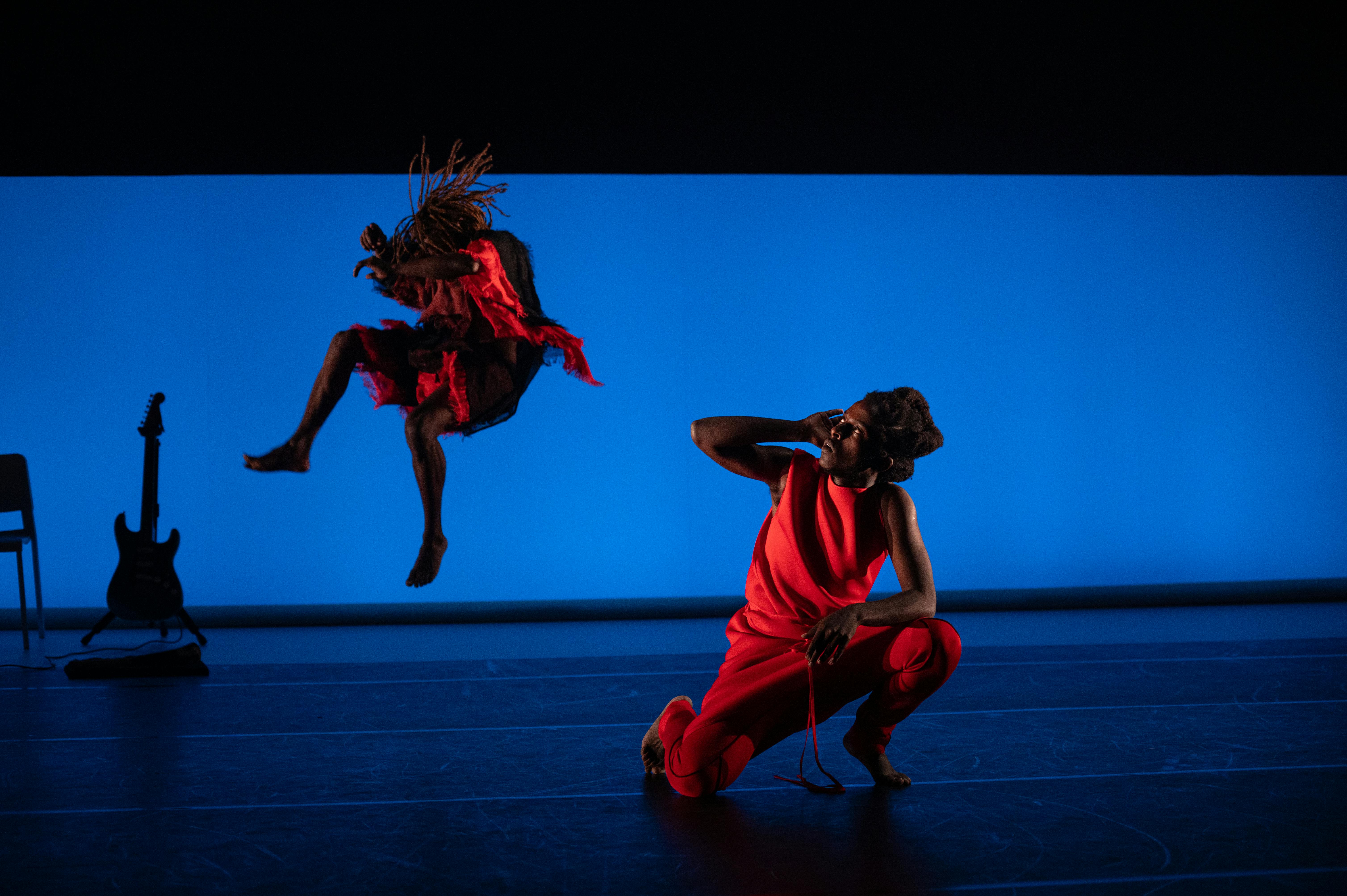 Two dancers on stage: one kneeling brings a hand to his ear while the second jumps