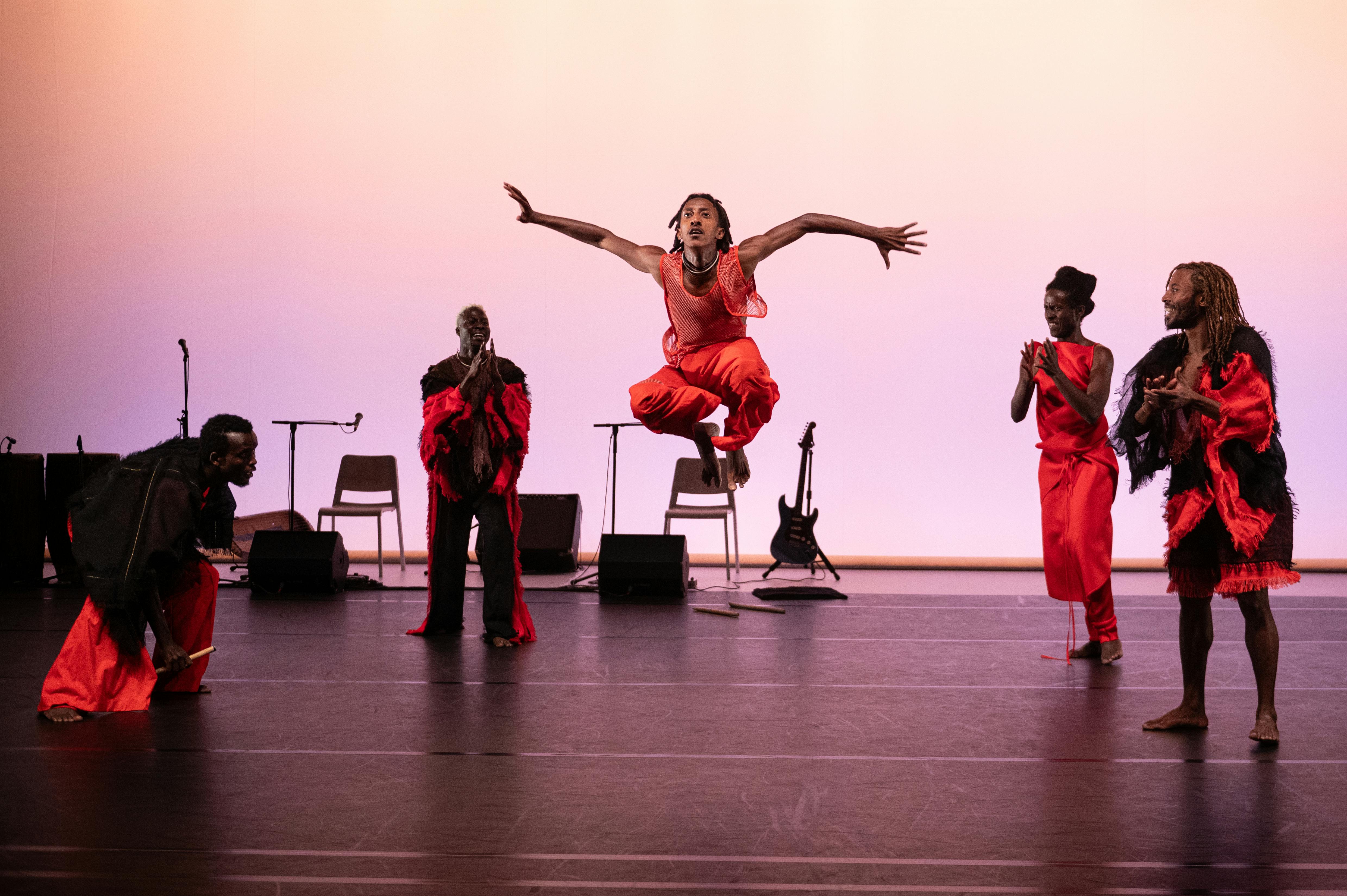 Five dancers on a red-lit stage stand in front of musical instruments
