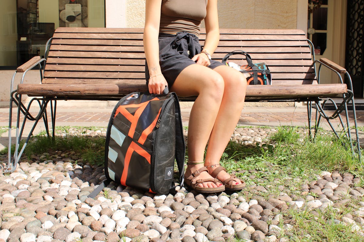 Outdoor photograph of a girl sitting on a bench with a backpack of our merchandising at her side