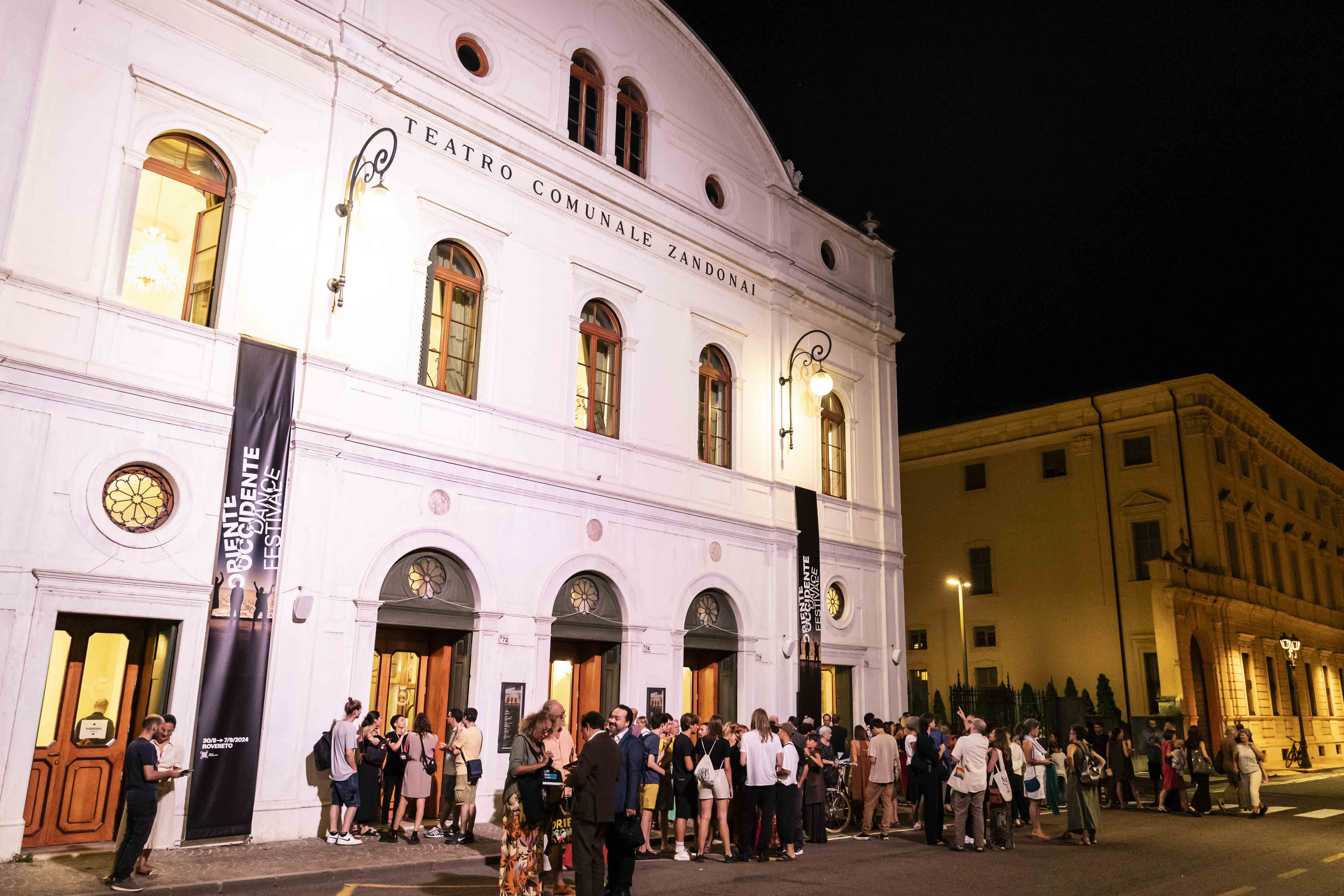 Un folto gruppo di persone è all'ingresso del teatro Zandonai di Rovereto. Sul teatro sono appesi i banner verticali di Oriente Occidente.
