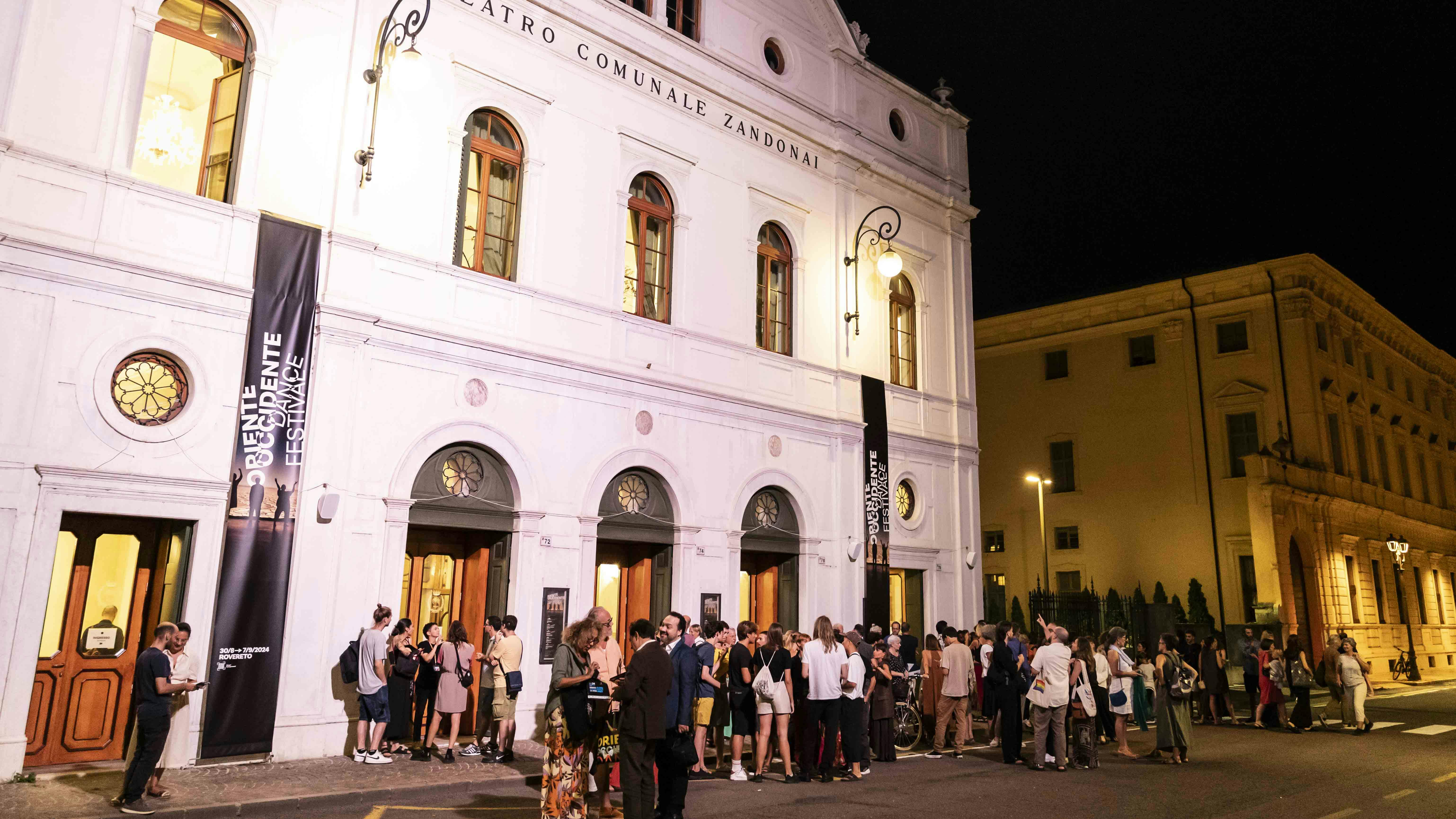 44° Oriente Occidente Dance Festival. Serata inaugurale al Teatro Zandonai Un folto gruppo di persone è all'ingresso del teatro Zandonai di Rovereto. Sul teatro sono appesi i banner verticali di Oriente Occidente.