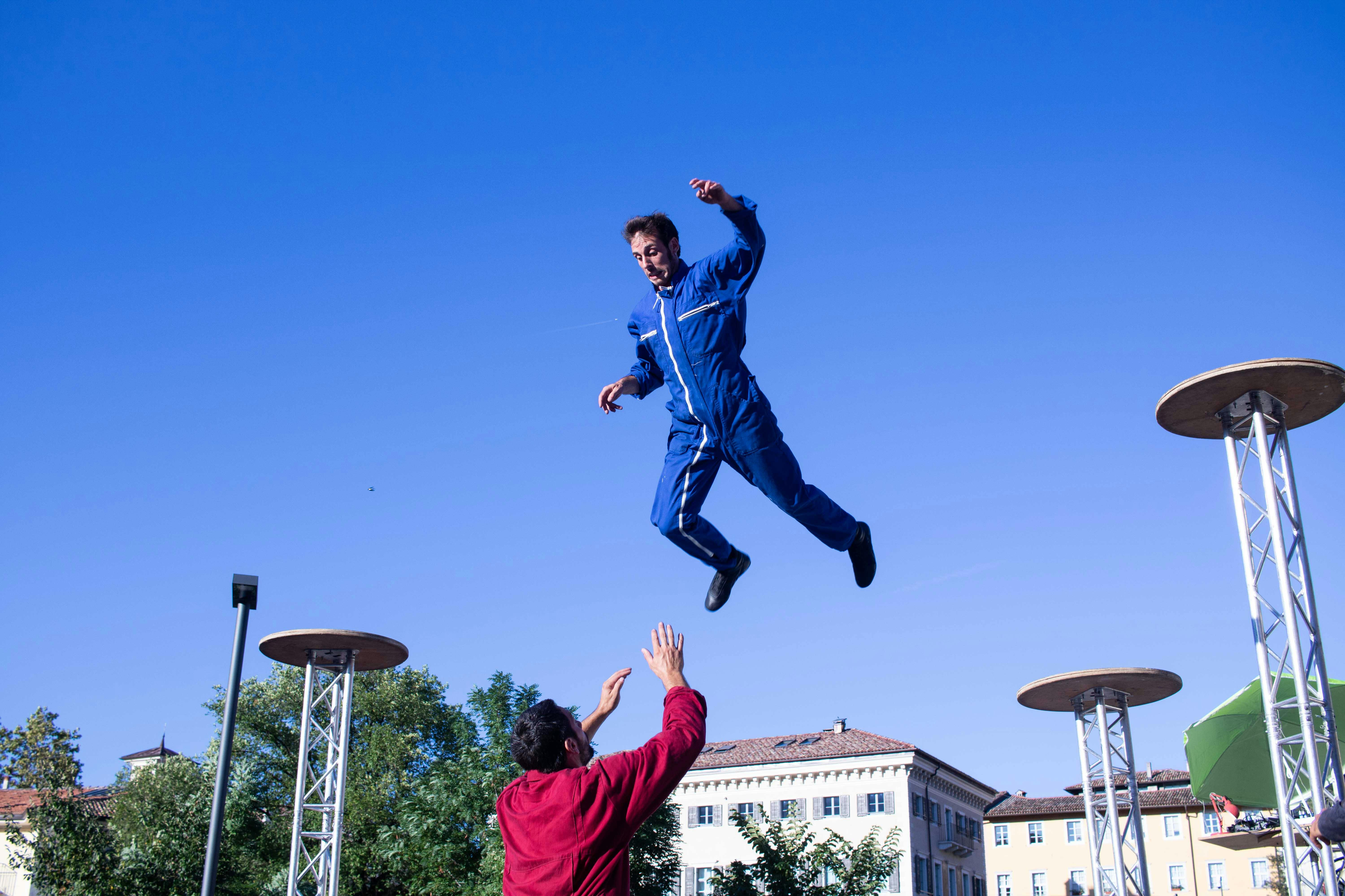 Un performer è in aria. Sotto di lui un altro gli tende le braccia. Sono in una piazza