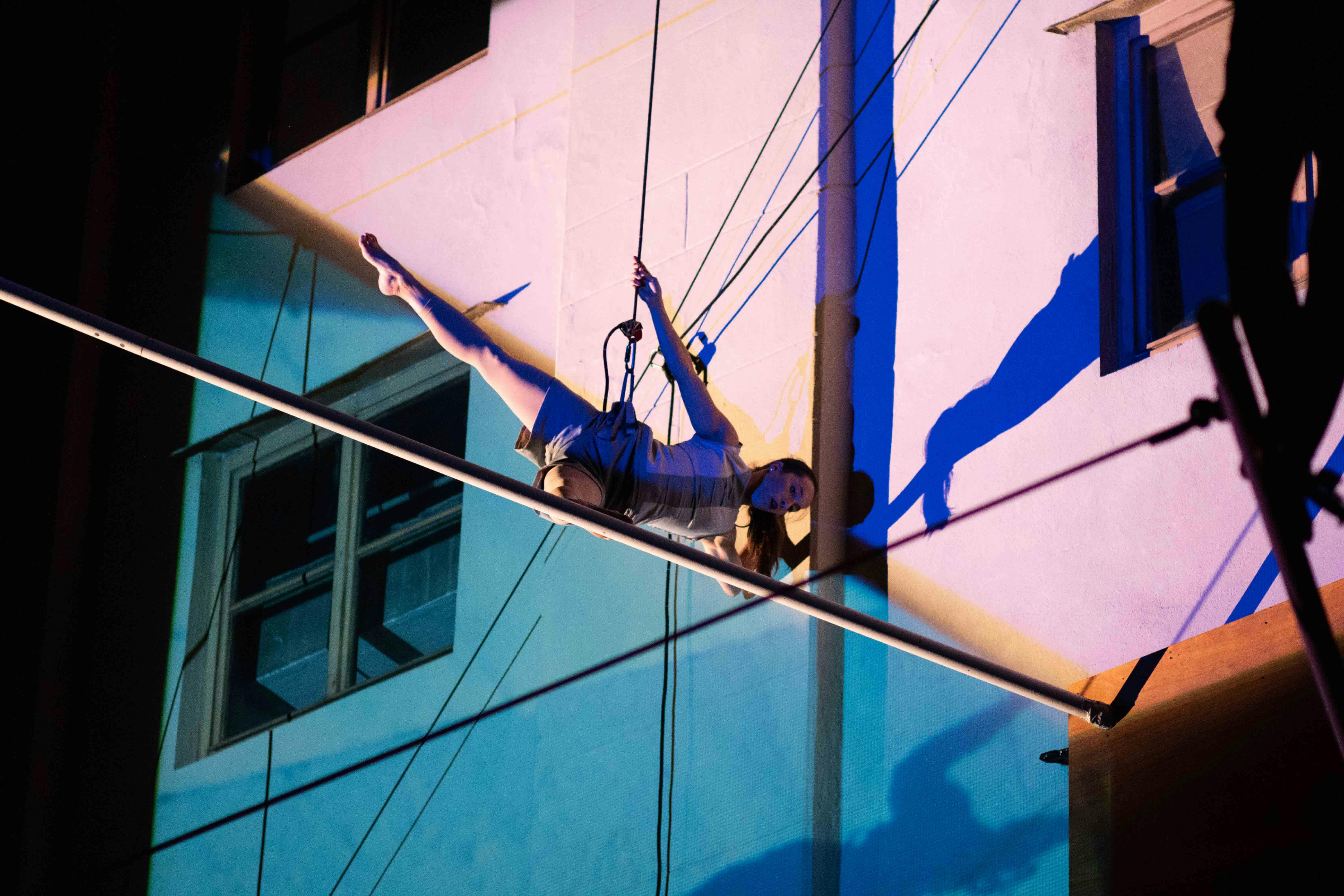 A performer hanging from ropes choreographs against the wall of a building at night