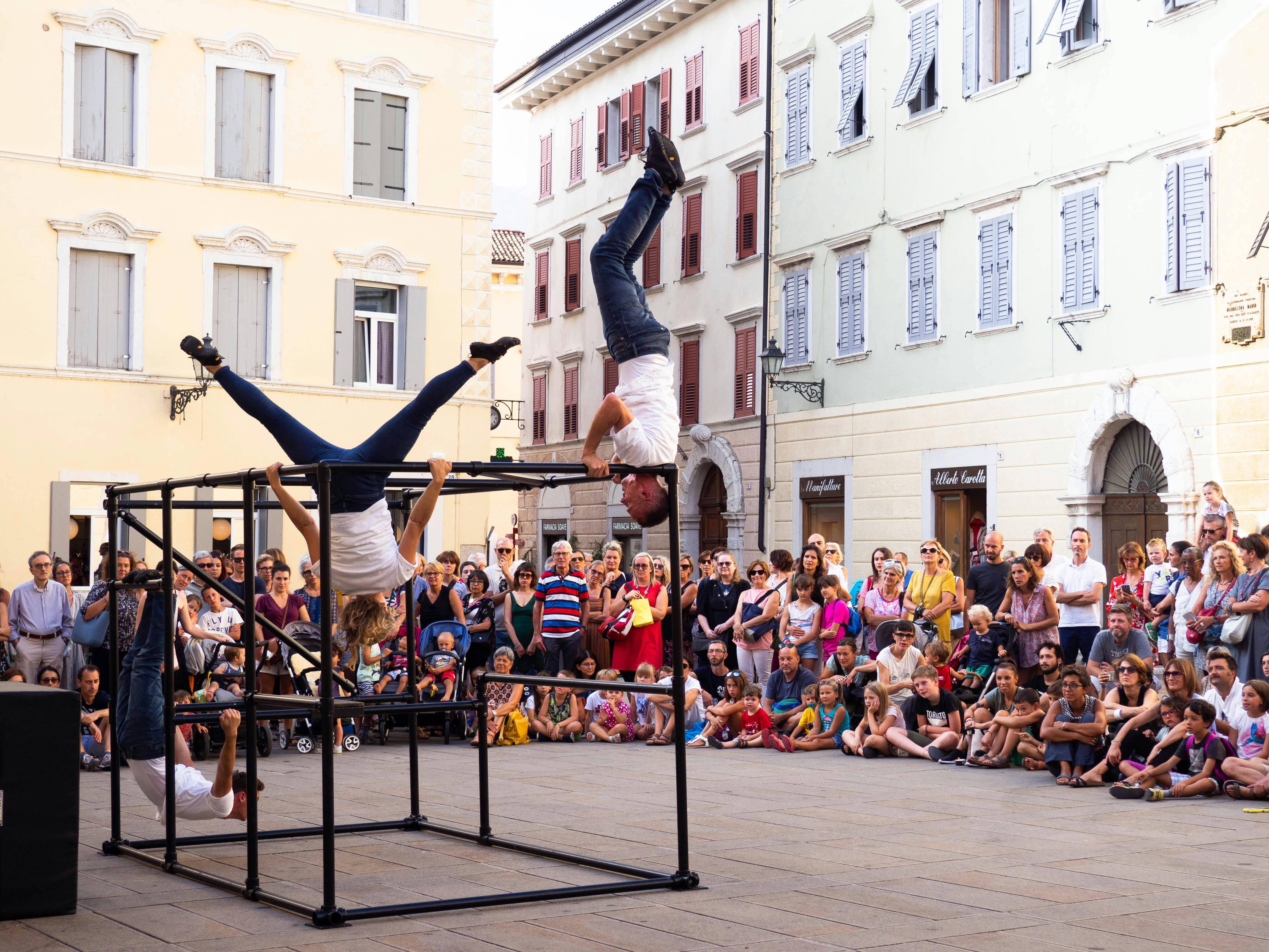 Three artists are gripping a cube-shaped structure made of poles. Surrounding them is a large audience seated on the ground.