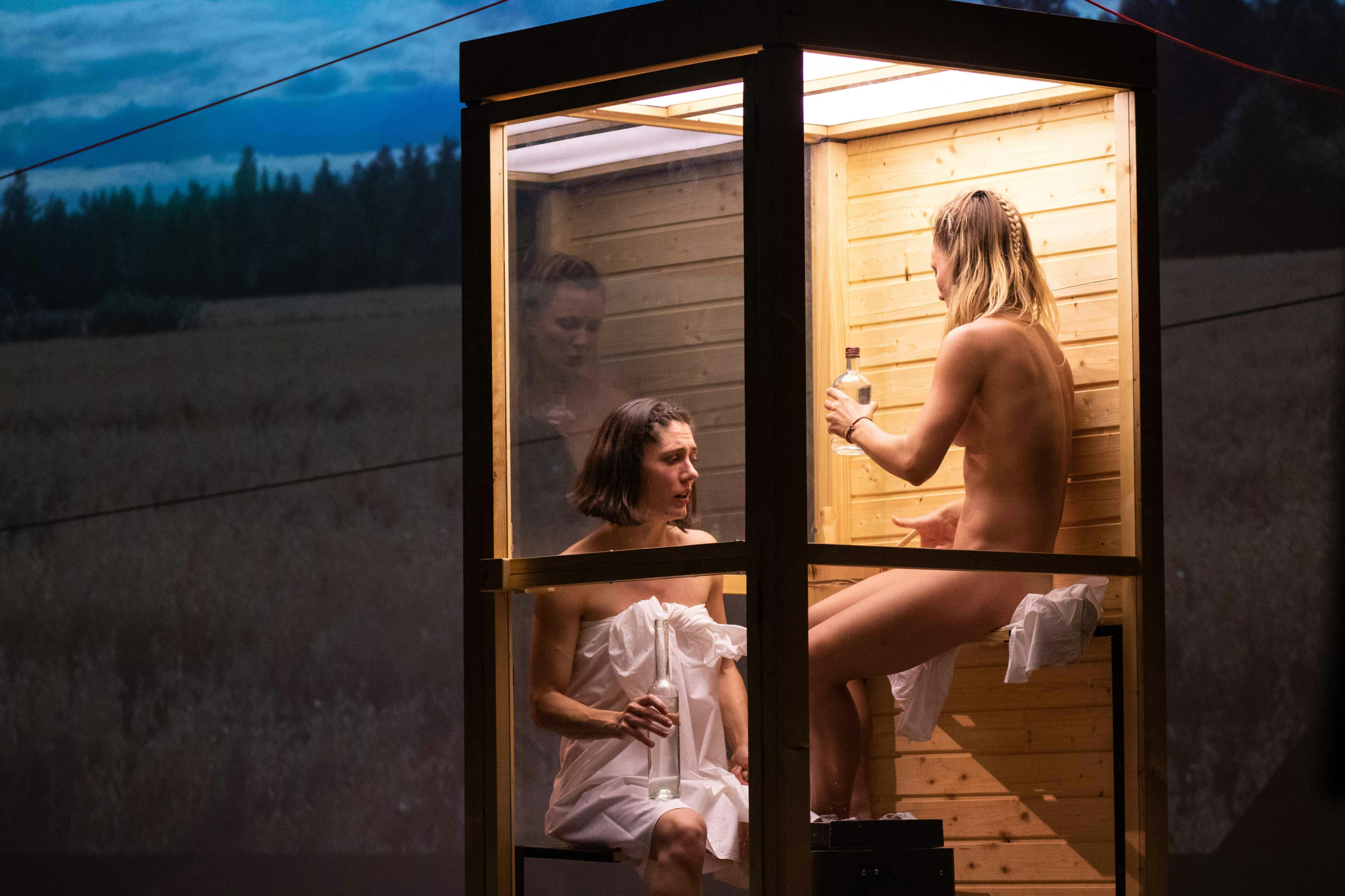 Two performers are inside a small sauna. One is without clothes and the other is wearing a towel. They are drinking and talking. A natural landscape is projected behind them