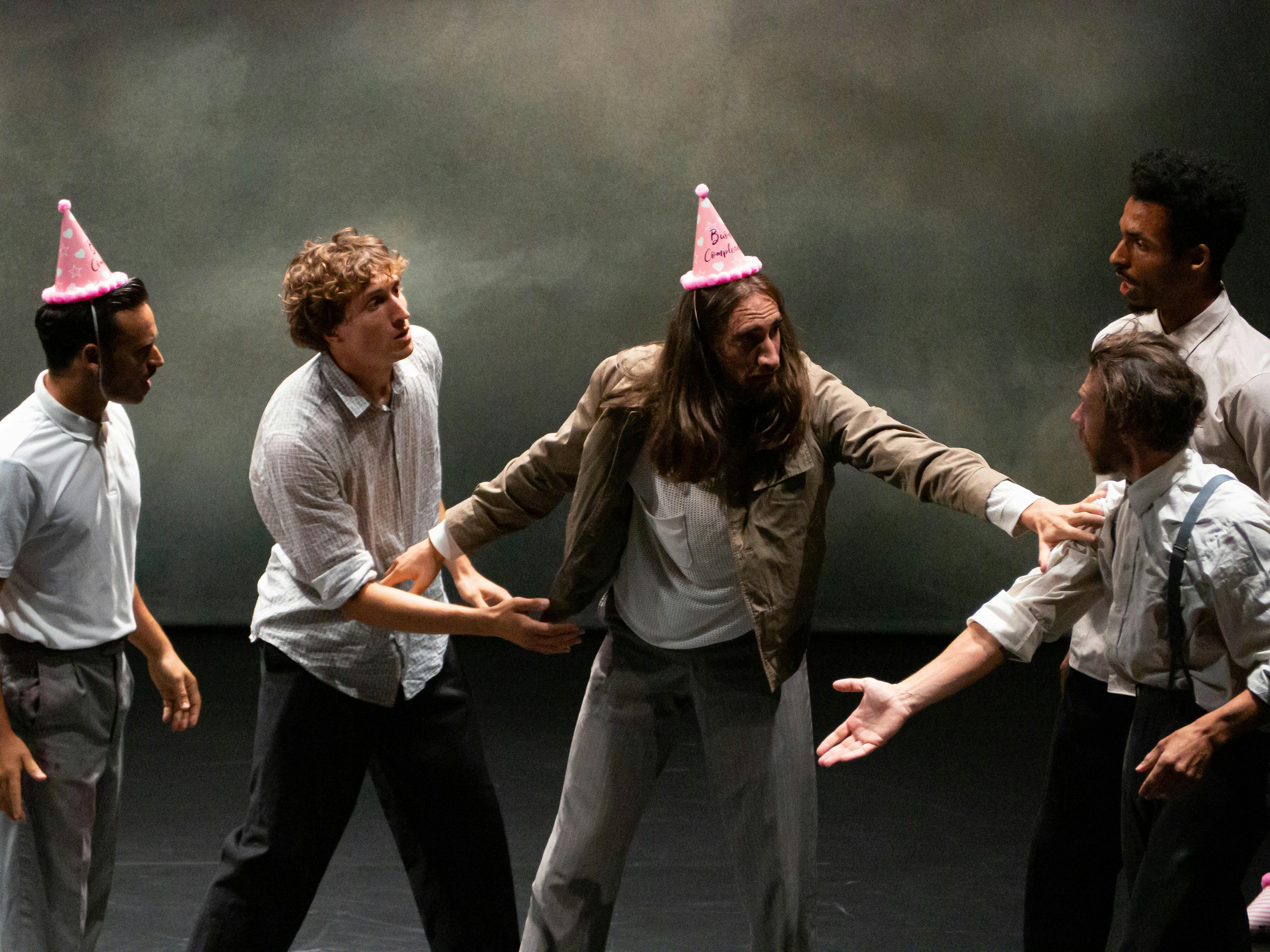 Un gruppo di performer sul palco indossano camice bianche. Uno al centro indossa un cappellino da compleanno e sta allontanando con le braccia gli altri