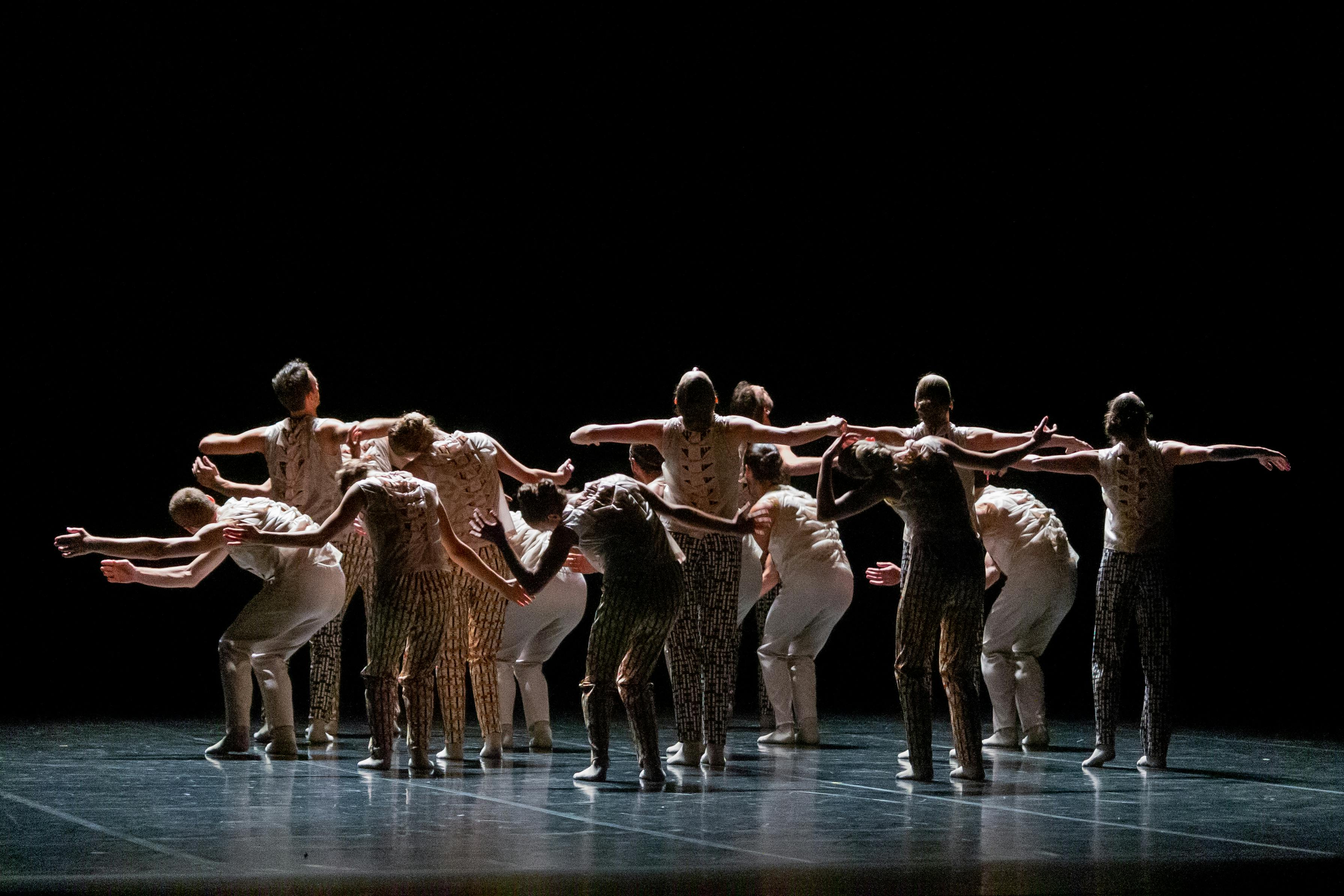 A group of performers on stage are photographed from behind. Some are bent over others hold their arms out and look up