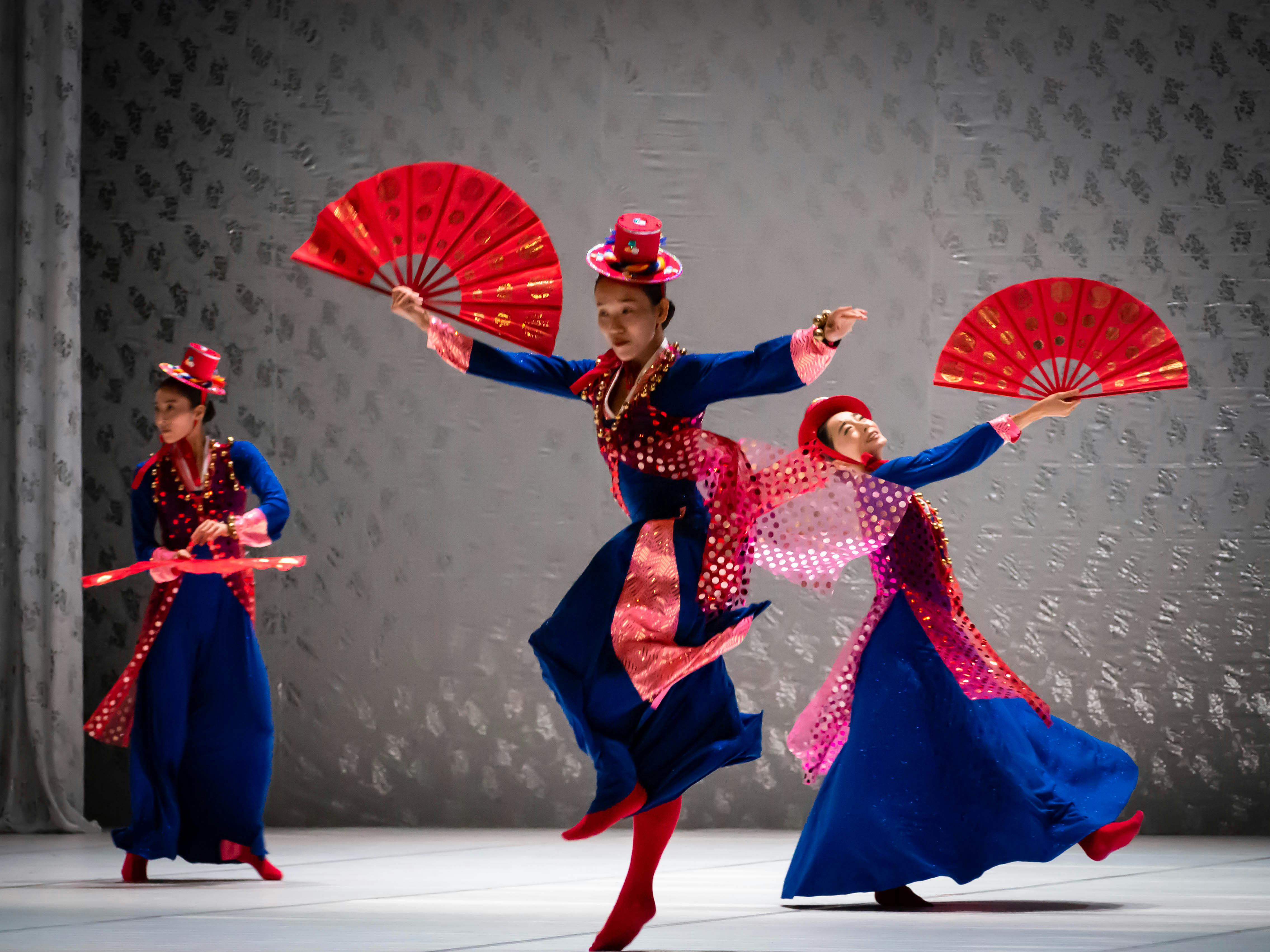 Three dancers are wearing blue and red dresses, wearing hats and holding large red fans