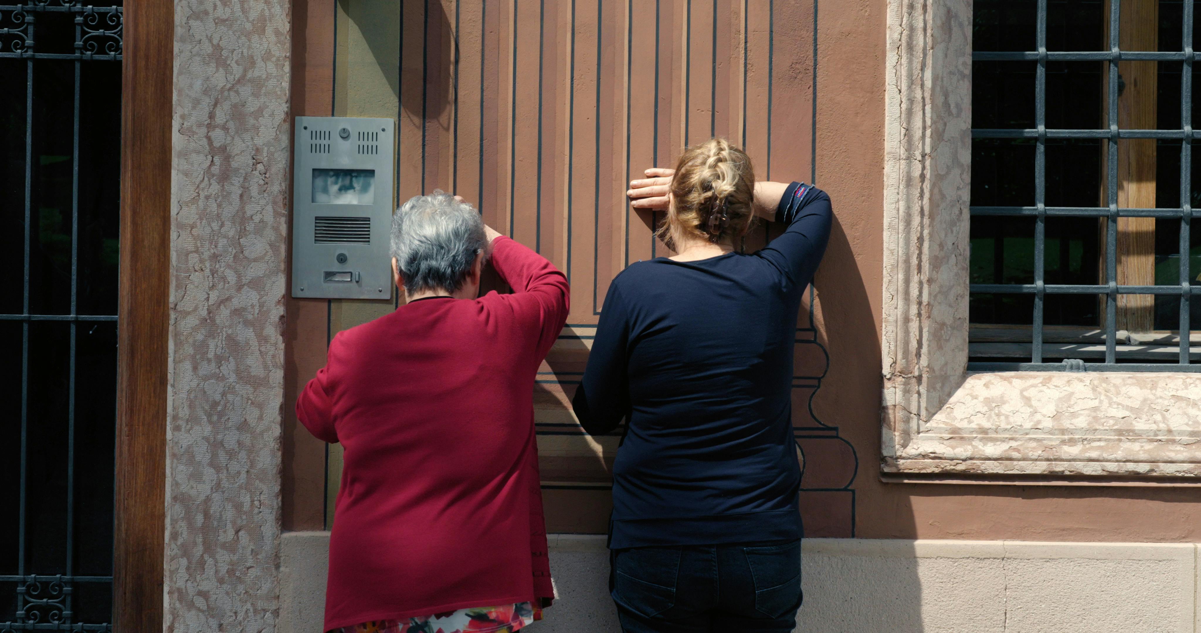 Due signore di schiena sono appoggiate a un muro con il braccio destro sollevato a coprire gli occhi