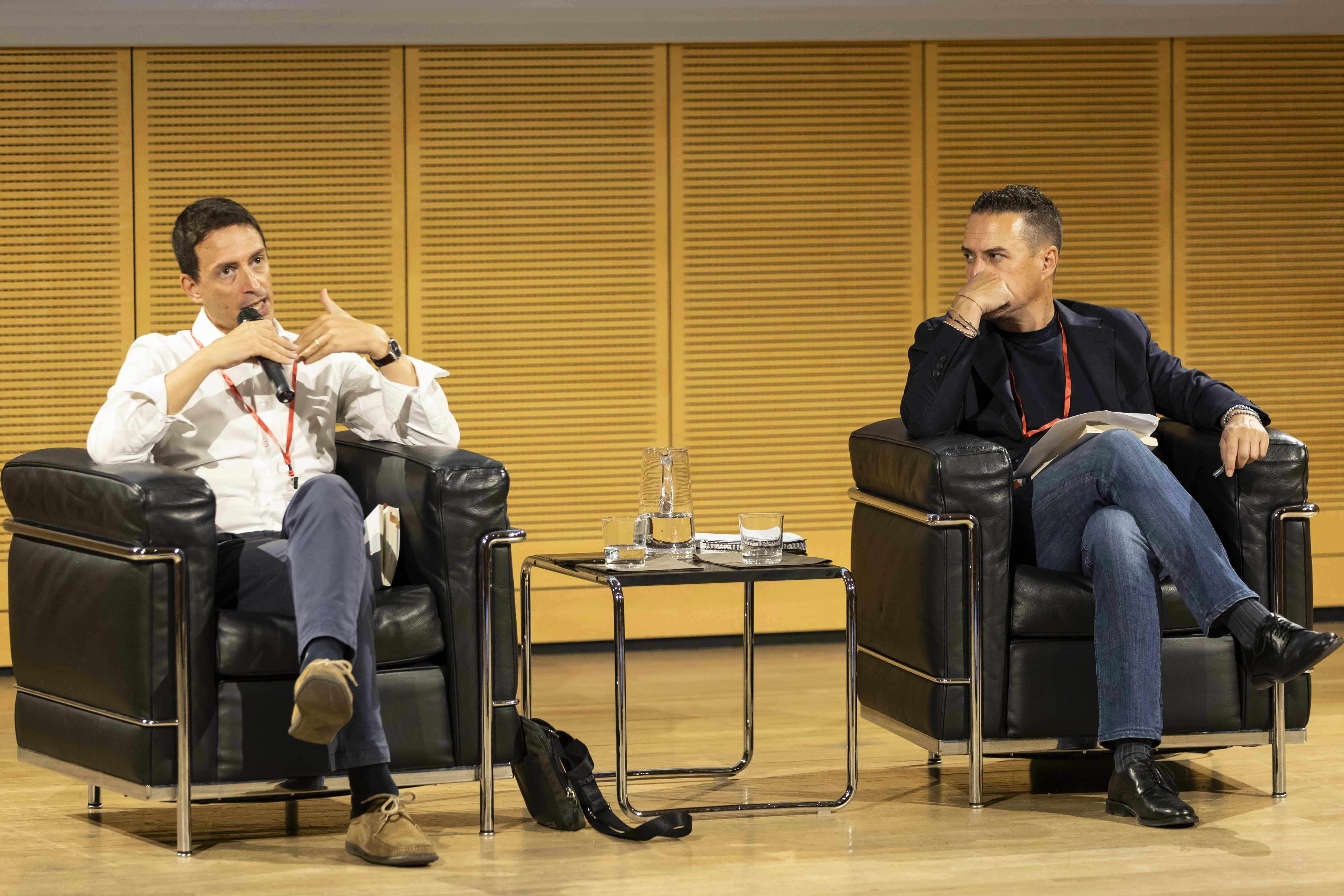 Foto di Fabrizio Maronta e Simone Casalini durante una conferenza. Sono entrambi sul palco seduti su delle poltrone nere. Maronta sta parlando al microfono.