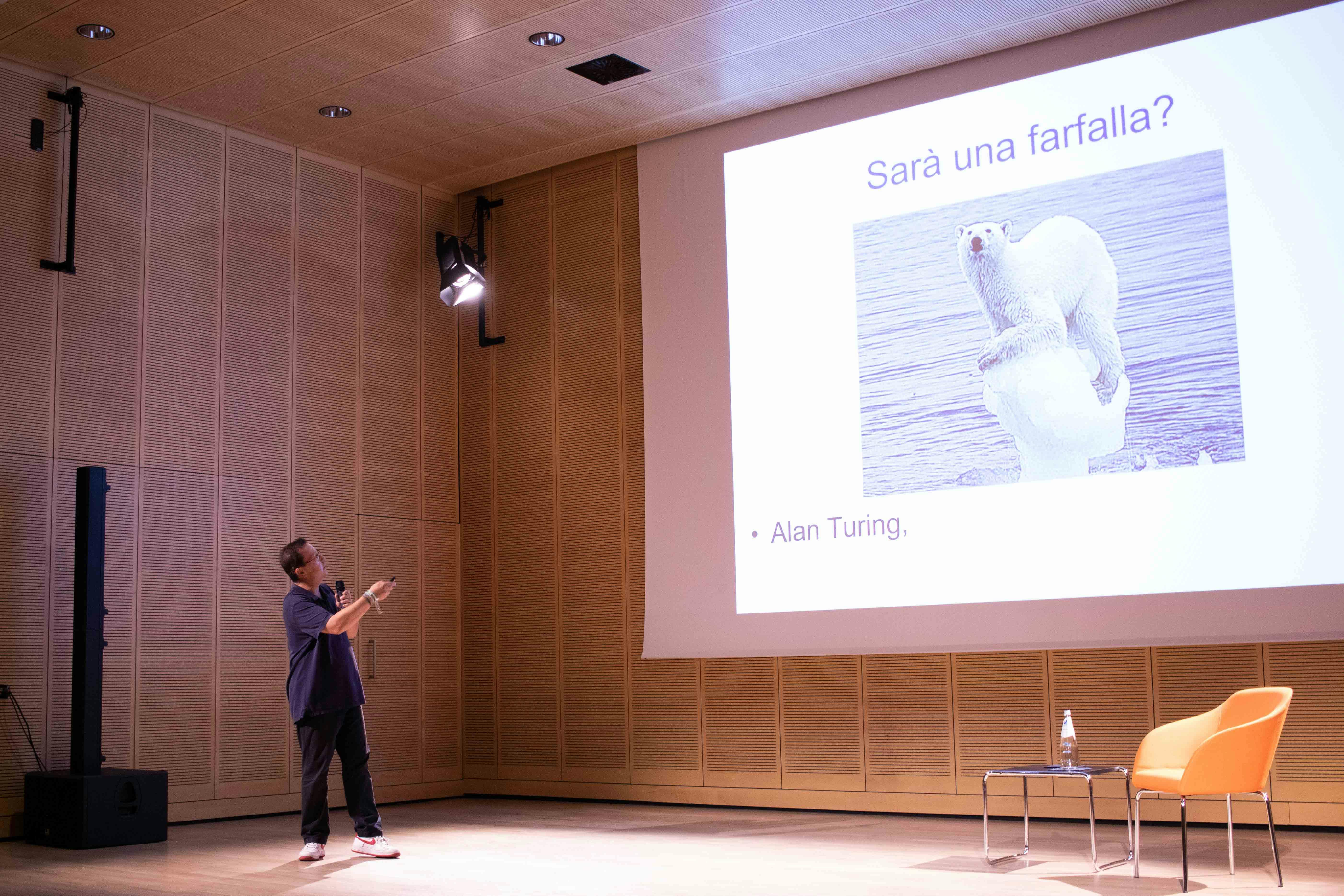 In foto Grammenos Mastrojeni è diplomatico, docente e scrittore sul palco. Indica la slide dietro di sè.