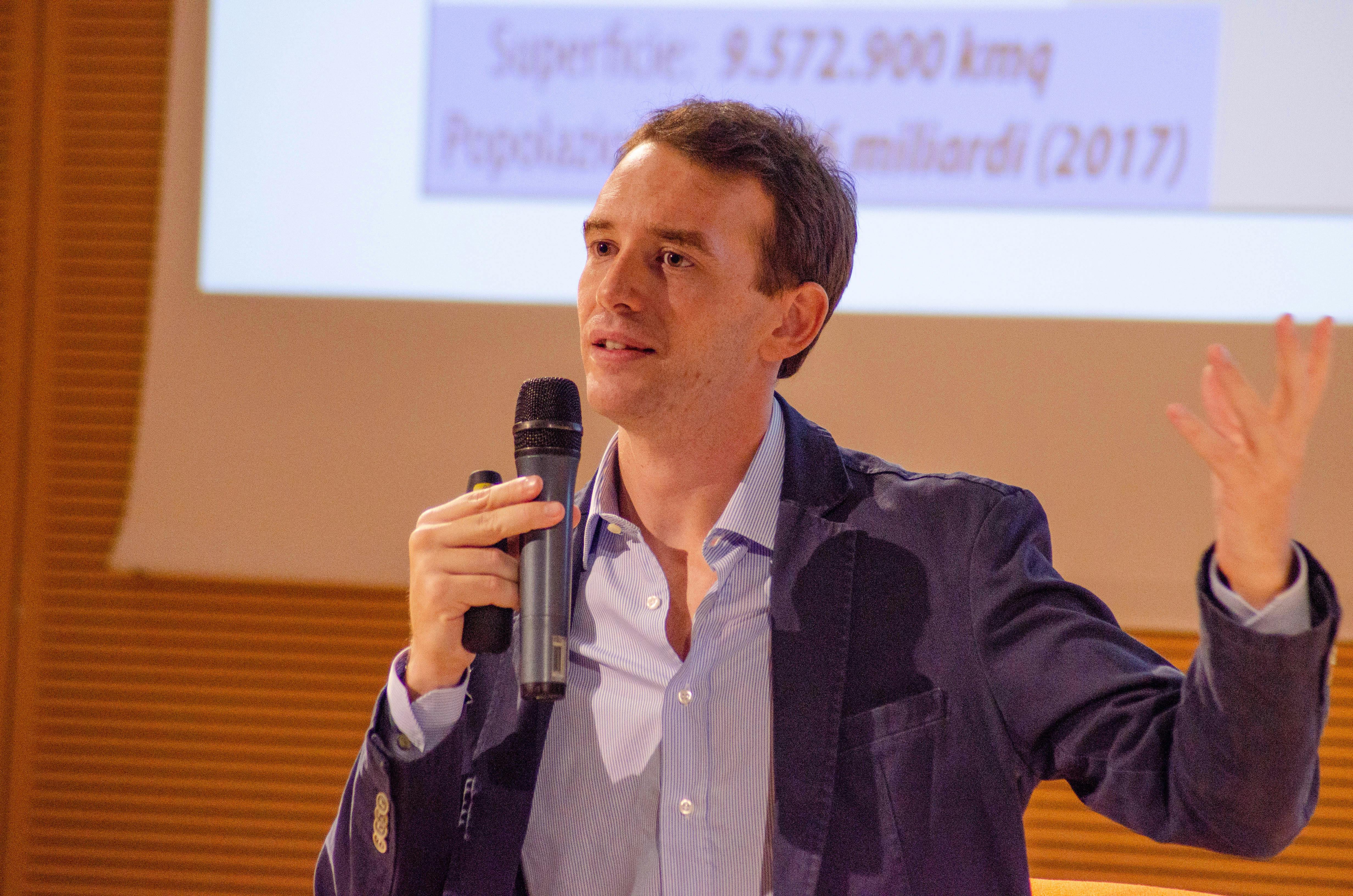 In foto Giorgio Cuscito, consigliere redazionale di Limes. è sul palco e parla al microfono. Indossa un completo blu e camicia azzurra.