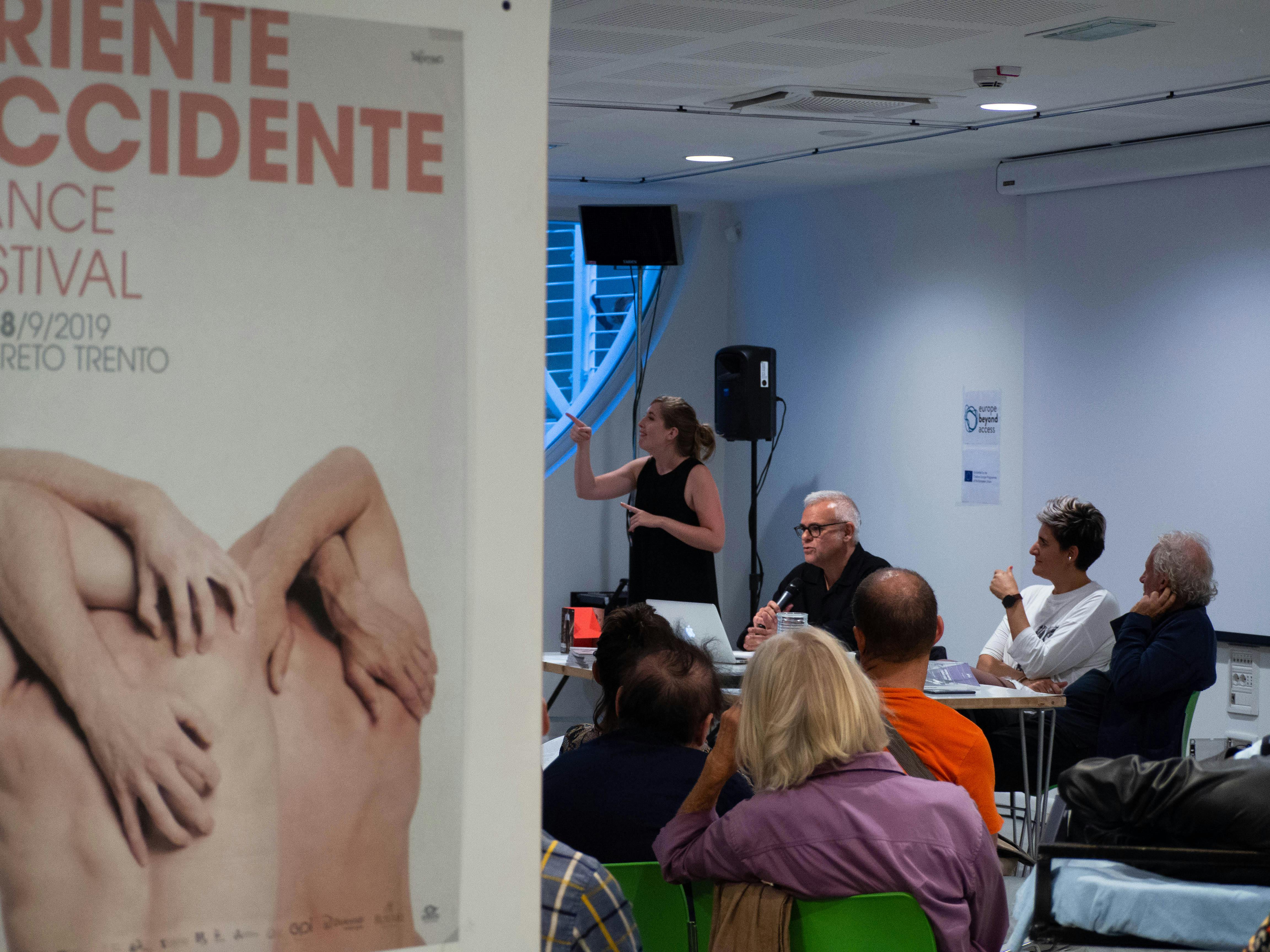 In primo piano sulla sinistra un poster di Oriente Occidente. Sullo sfondo tre persone al tavolo che parlano a una platea di persone.