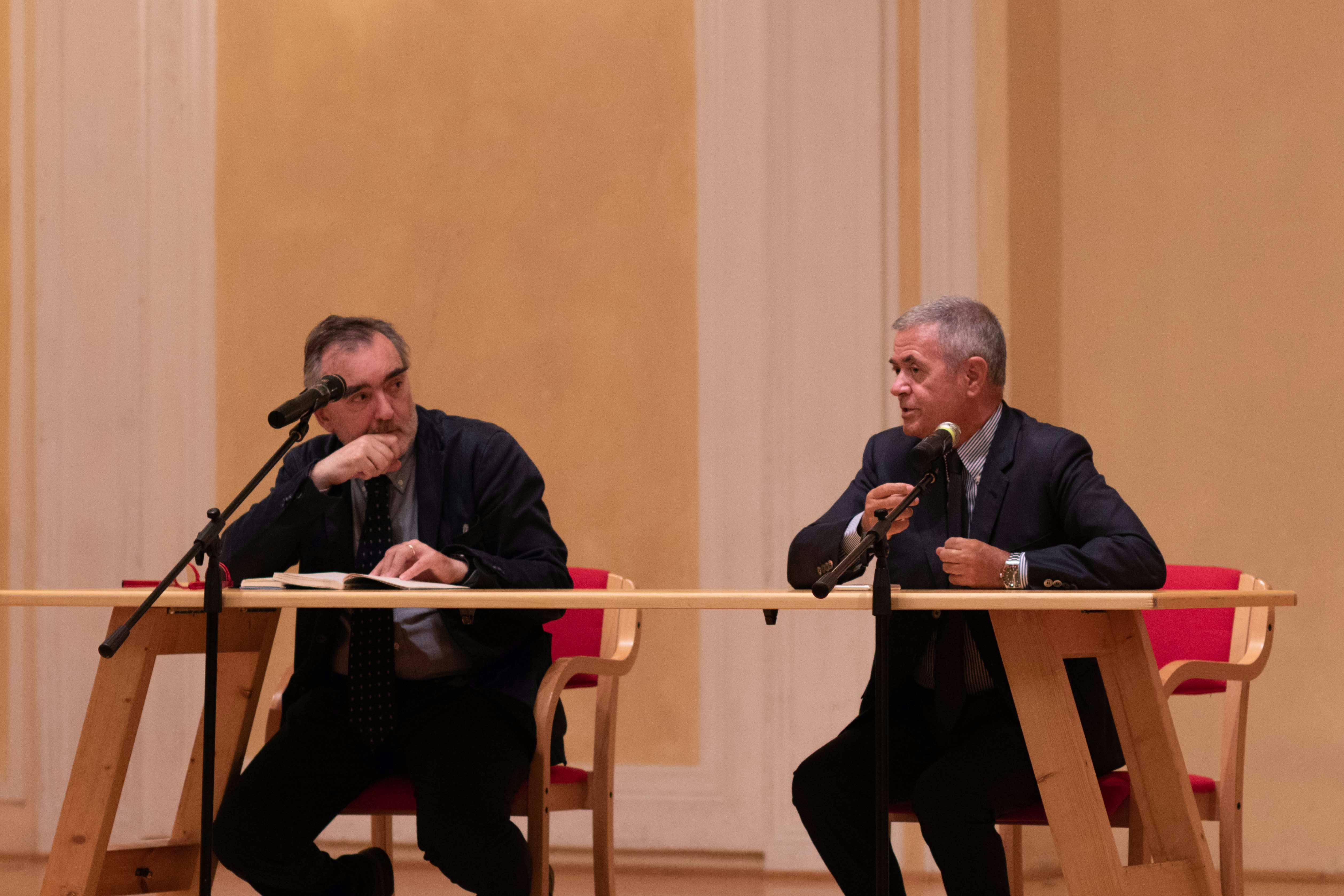 In the photo, Ezio Mauro and Alberto Faustini are sitting at a table with two microphones. Both are wearing dark suits.