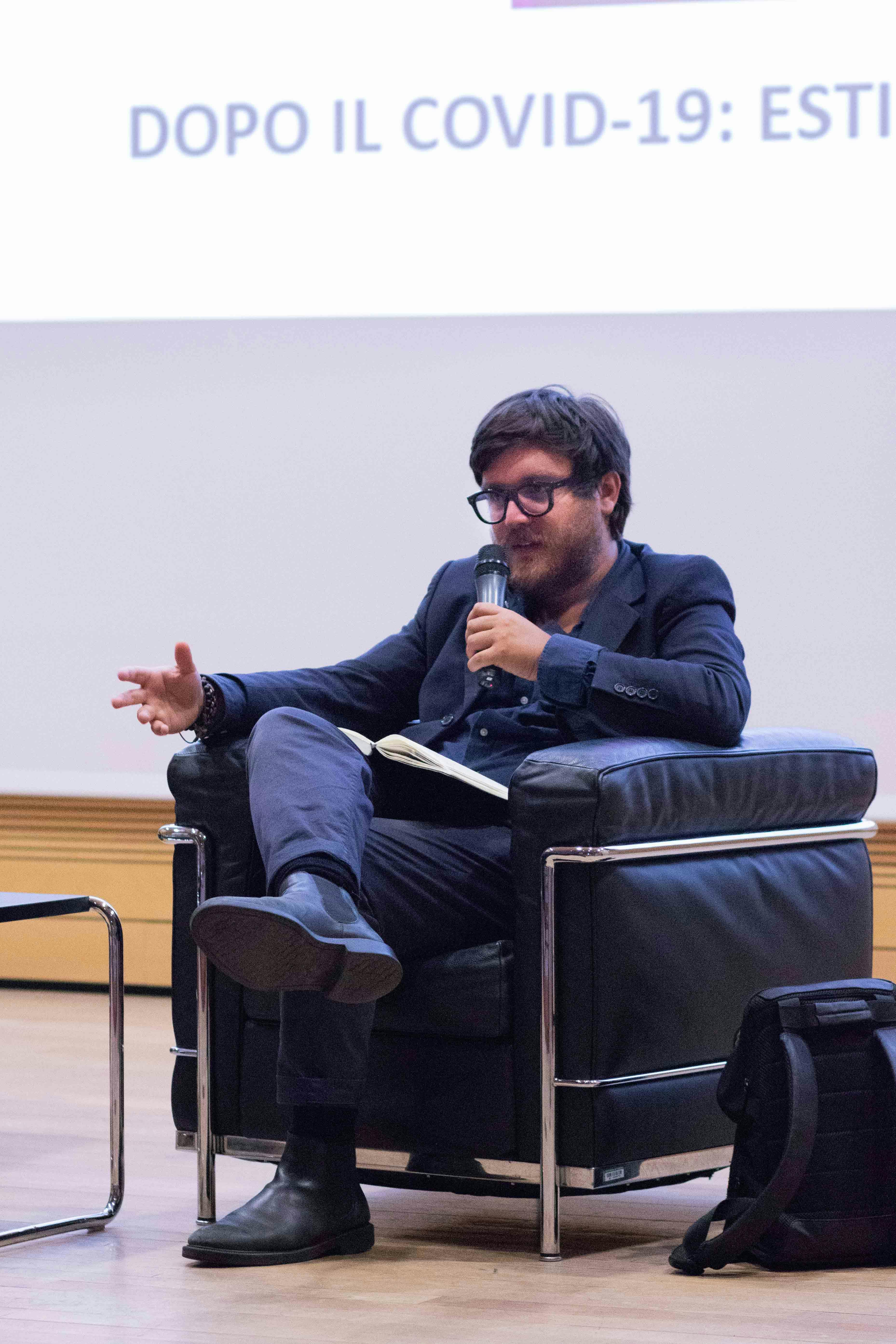 Leonardo Caffo on stage, sitting in an armchair. He is speaking into a microphone and wearing a blue suit.