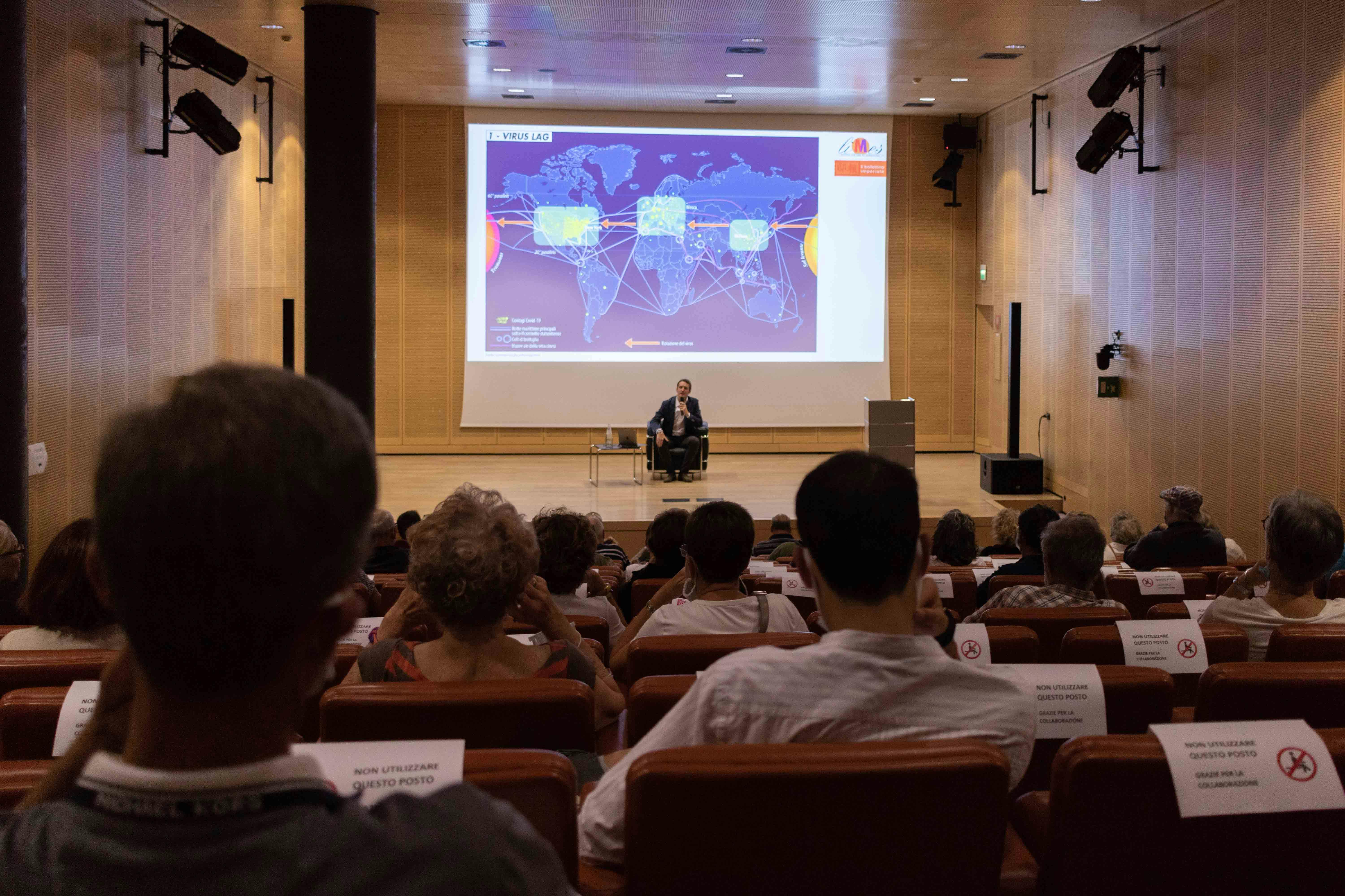 Audience with their backs to the camera, watching the stage where Giorgio Cuscito is seated.
