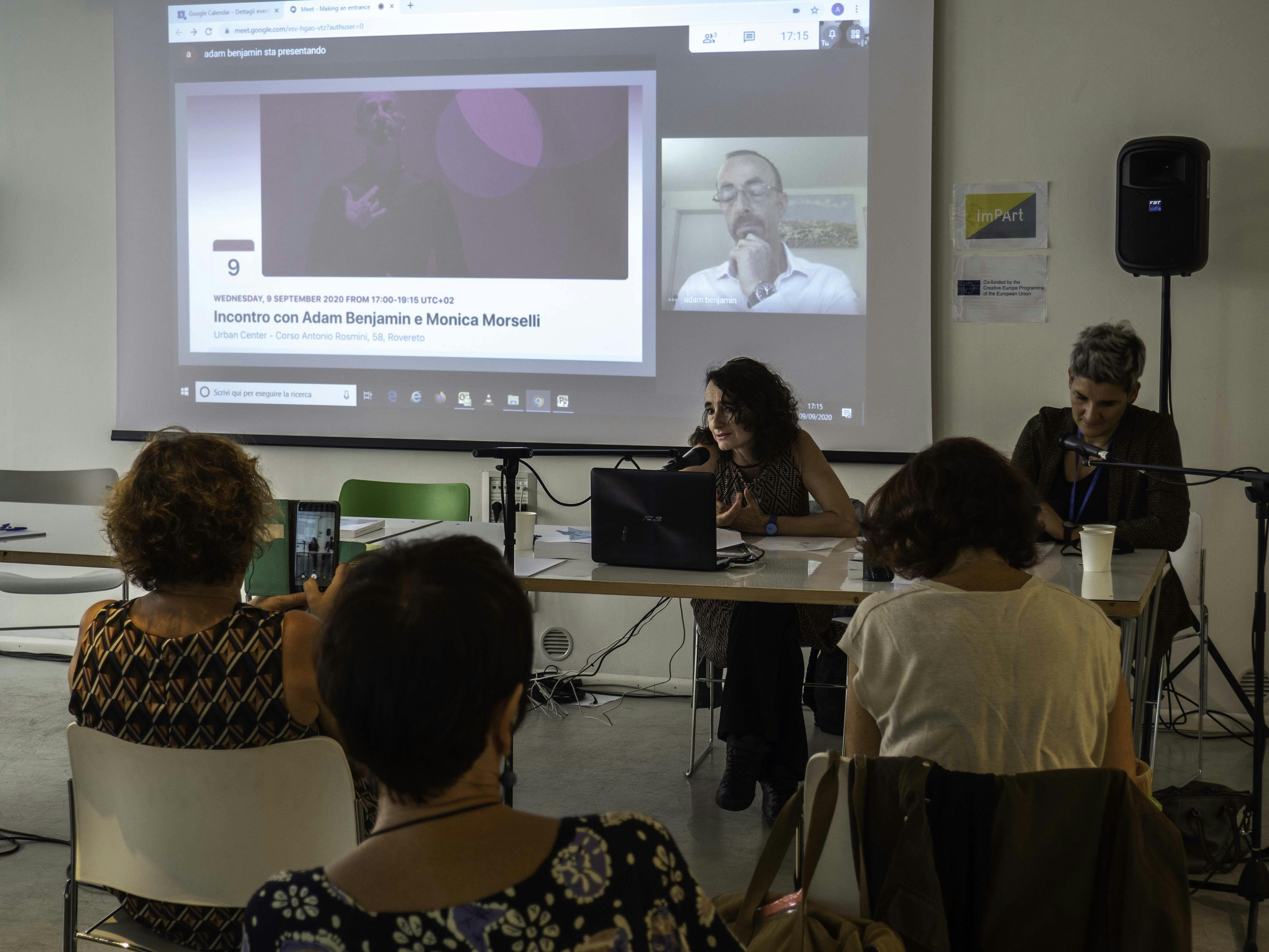 Foto di tre persone di spalle. Guardano due donne alla scrivania che parlano al microfono.