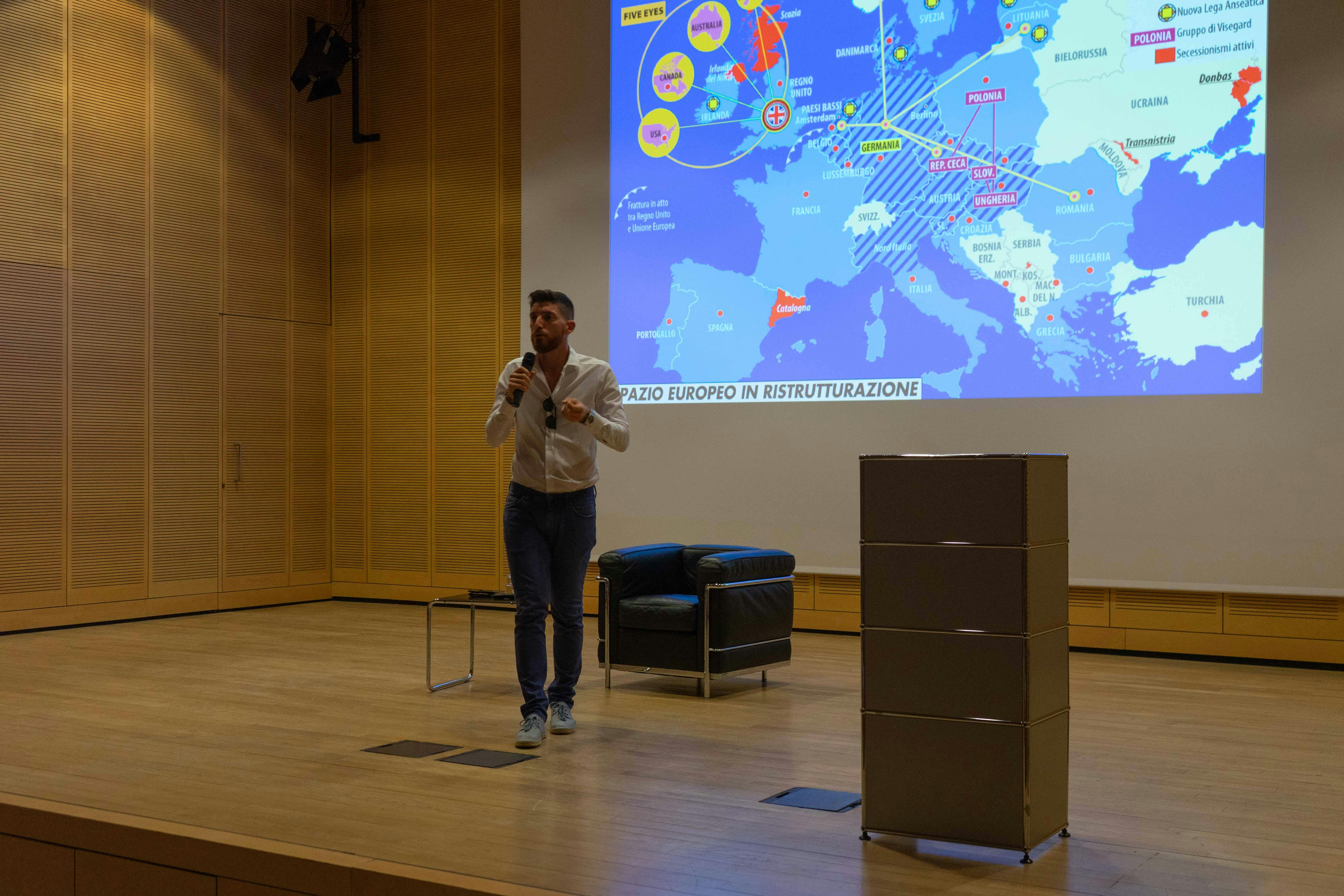 Sul palco Federico Petroni mentre parla al microfono. è in piedi. Dietro di lui è proiettata una mappa geografica con il proiettore.