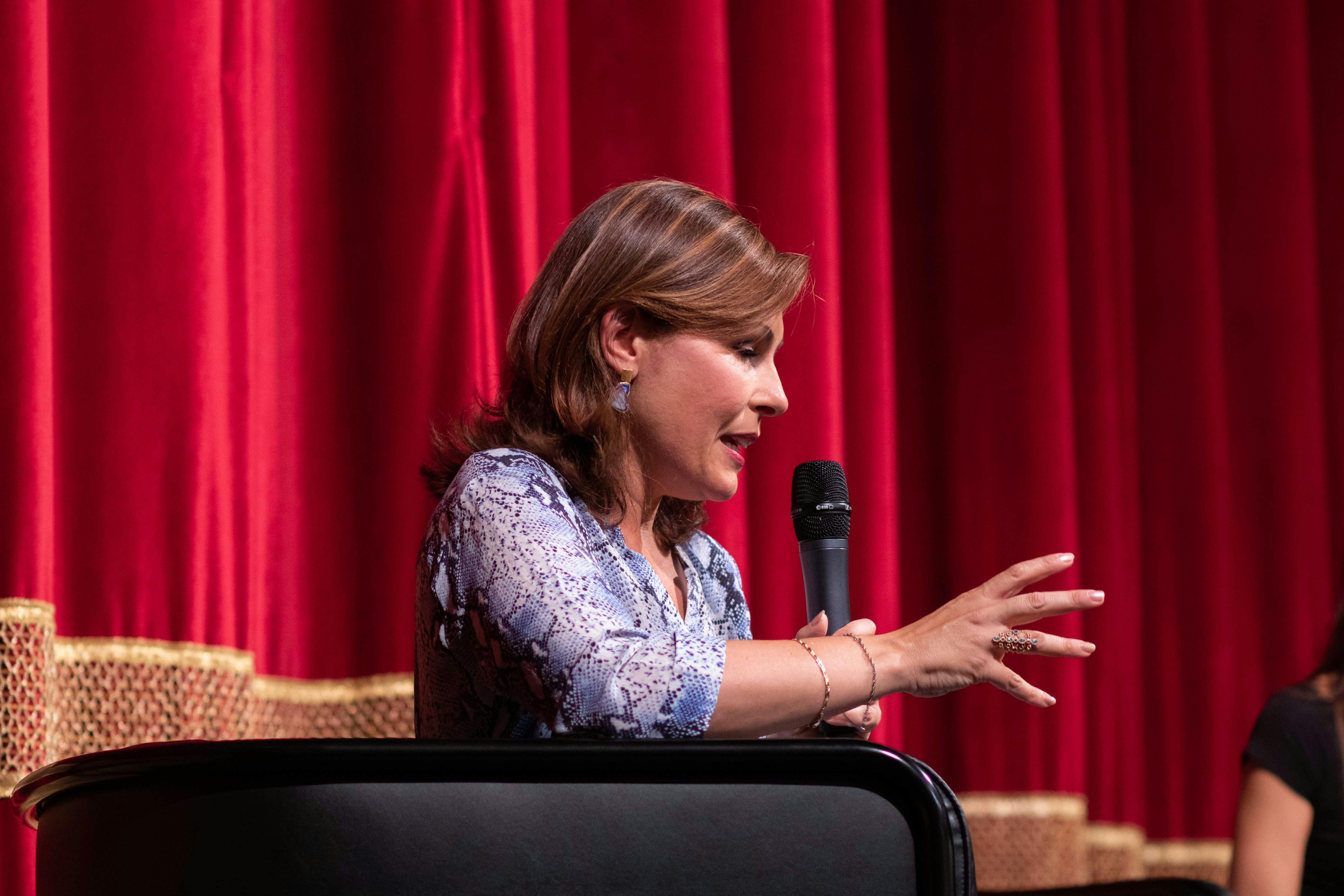 In foto Barbara Serra, giornalista. è seduta, parla al microfono. Sullo sfondo il sipario rosso del teatro.