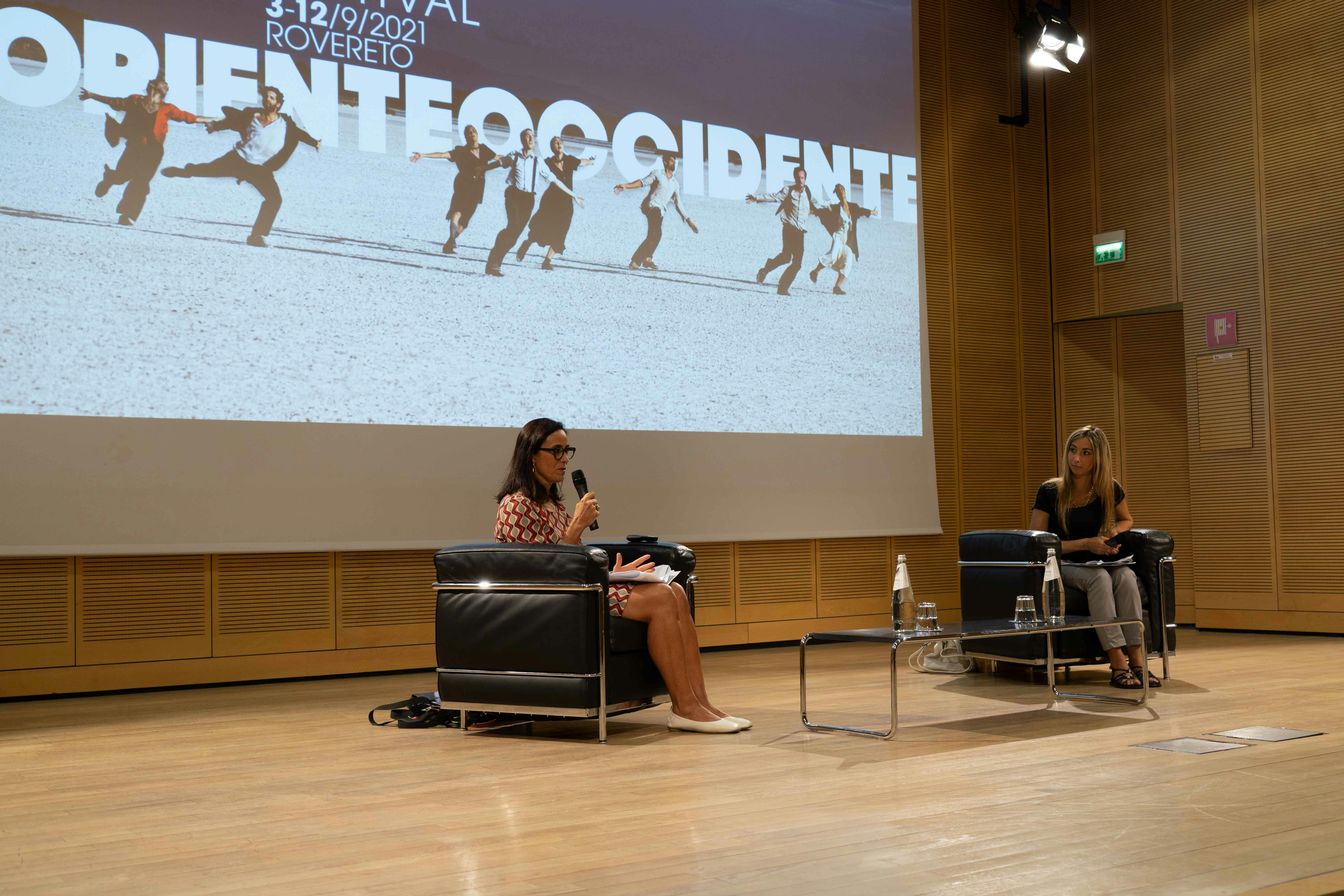 In the photo, Laura Canali is on stage speaking into a microphone while seated in an armchair. Another woman is on stage with her.