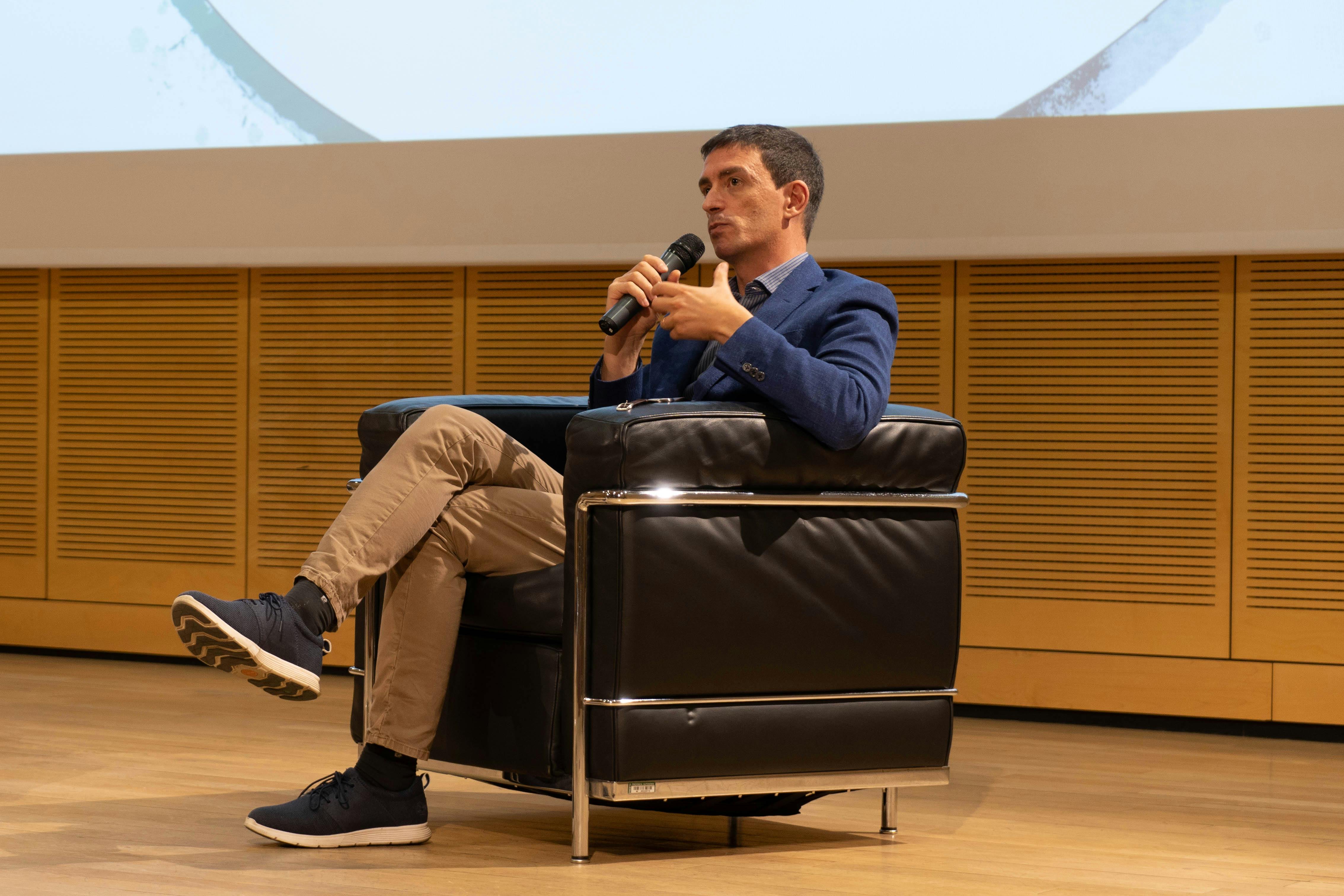 In foto Fabrizio Maronata sul palco mentre parla al microfono. è seduto su una poltrona scura.