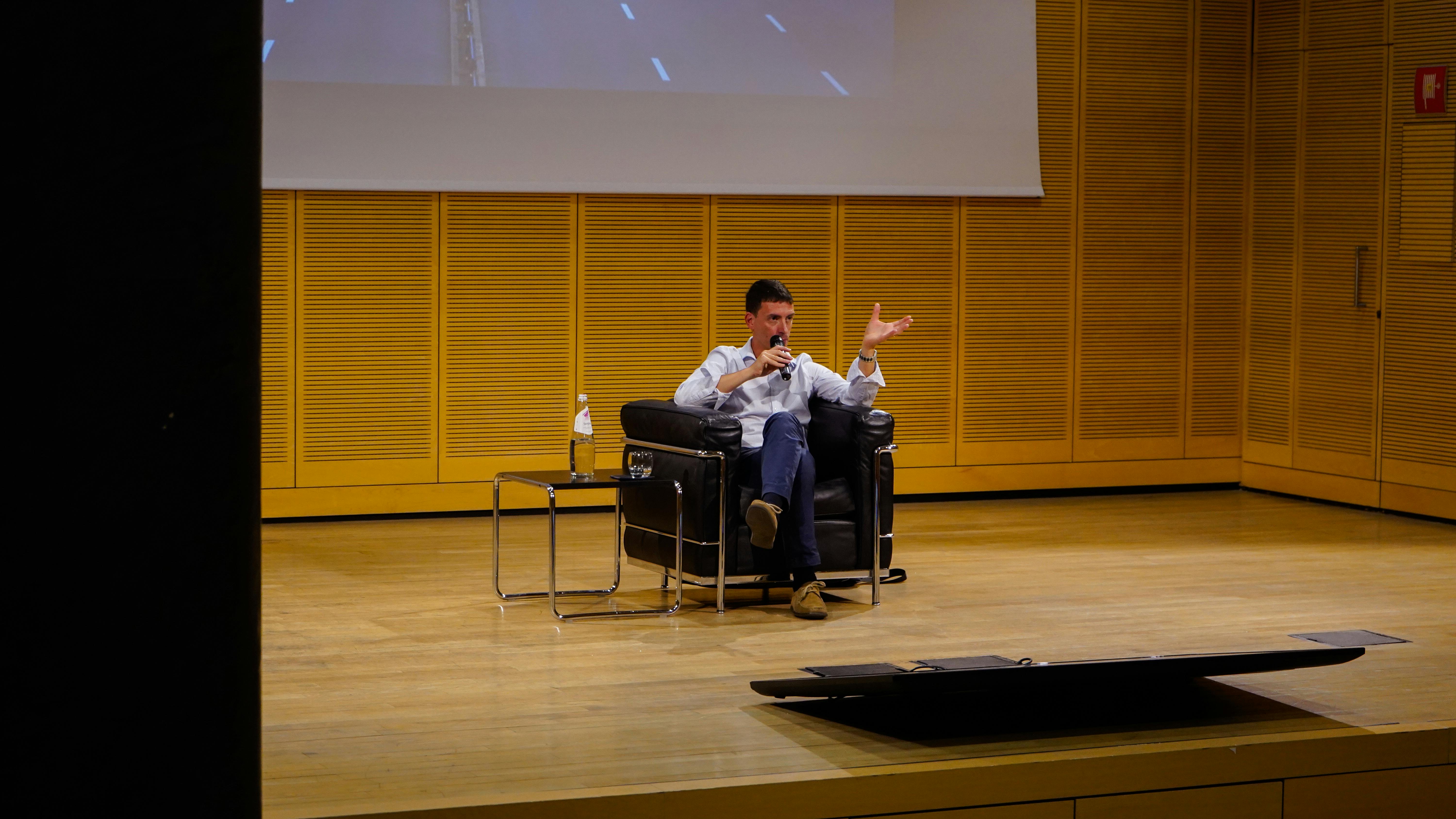 Foto di Fabrizio Maronta sul palco durante una conferenza. è seduto su una poltrona e sta parlando al microfono.