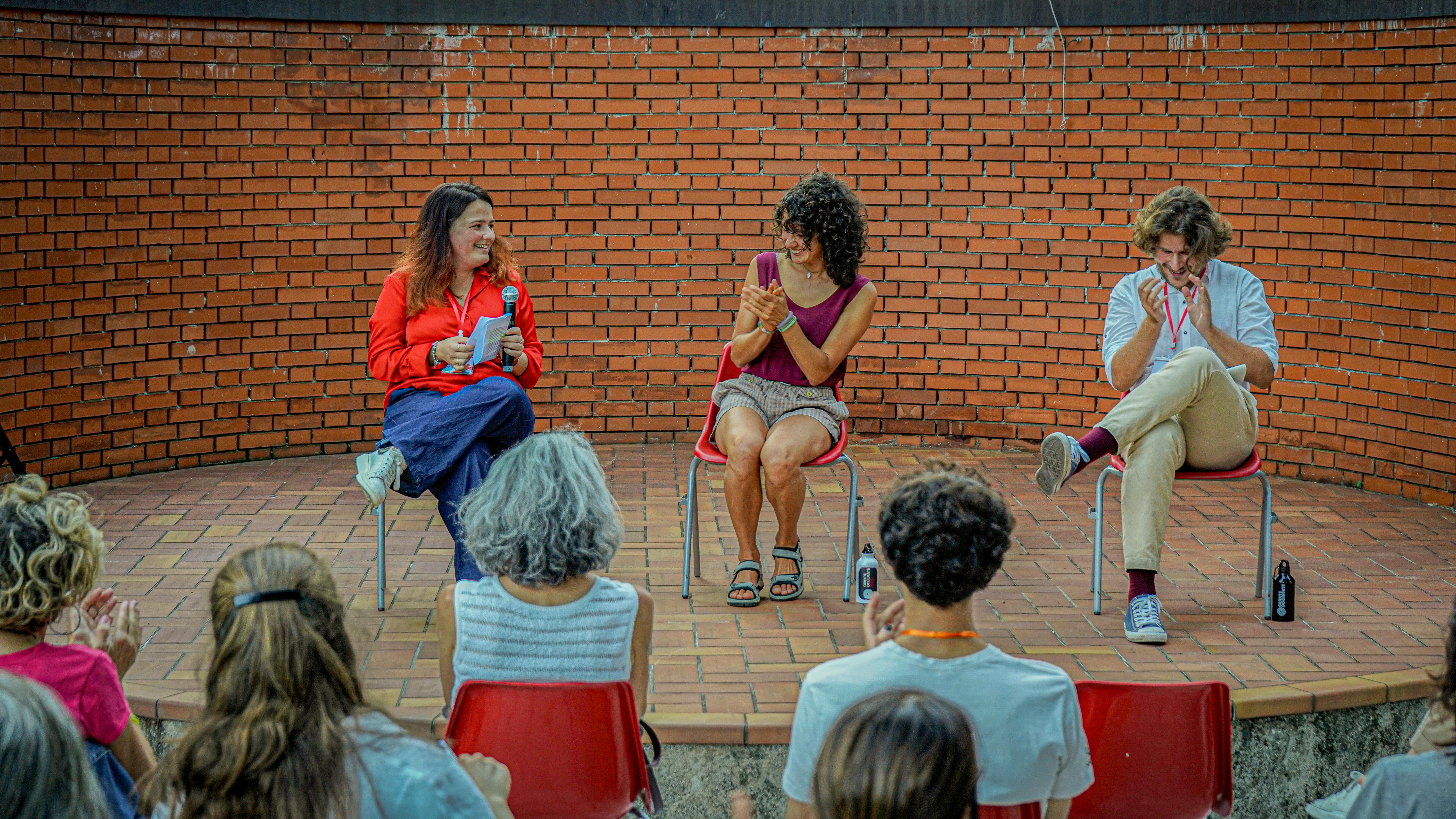 Sul palco all'aperto Sara Segantin, Silvia Kasper e Marzio Fait. Sono seduti su delle sedie.
