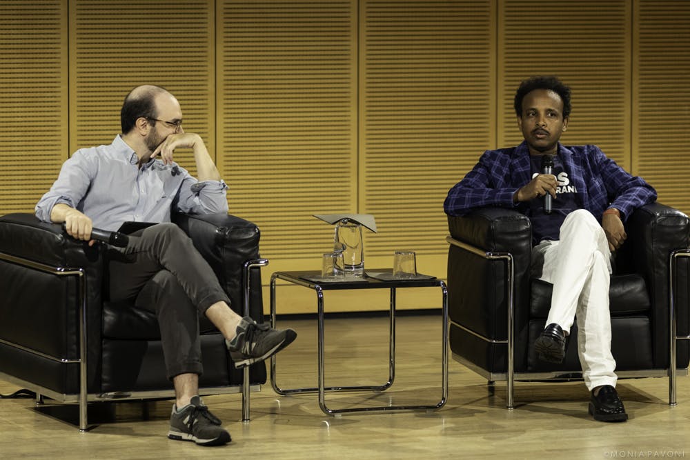 Foto di Abdelfetah Mohamed e Luca Misculin durante una conferenza. Sono entrambi sul palco seduti su delle poltrone nere. Mohamed sta parlando al microfono.