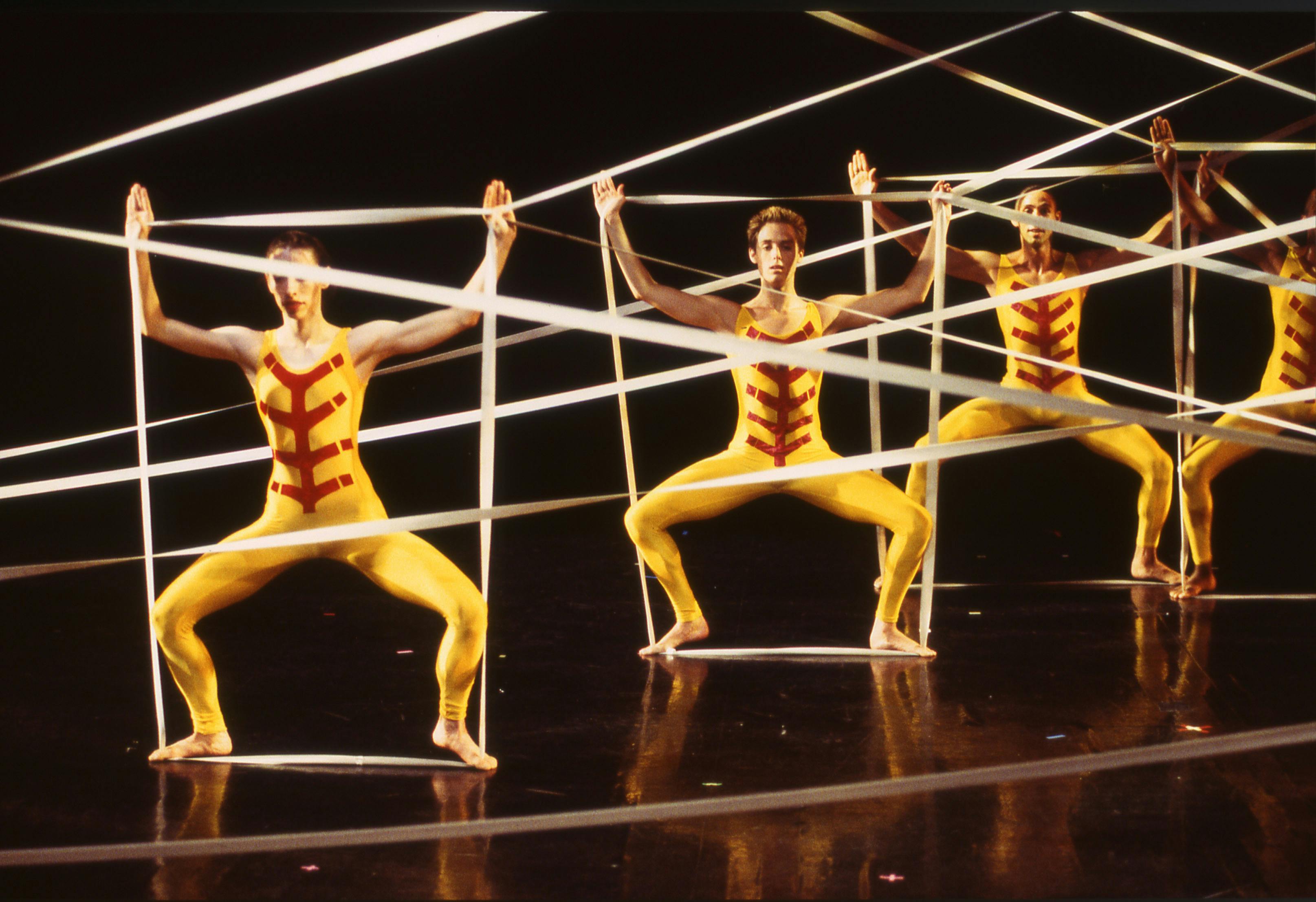 Four artists, dressed in yellow suits. Their arms and legs are spread wide as they hold a taut ribbon with their hands and feet to form a square.