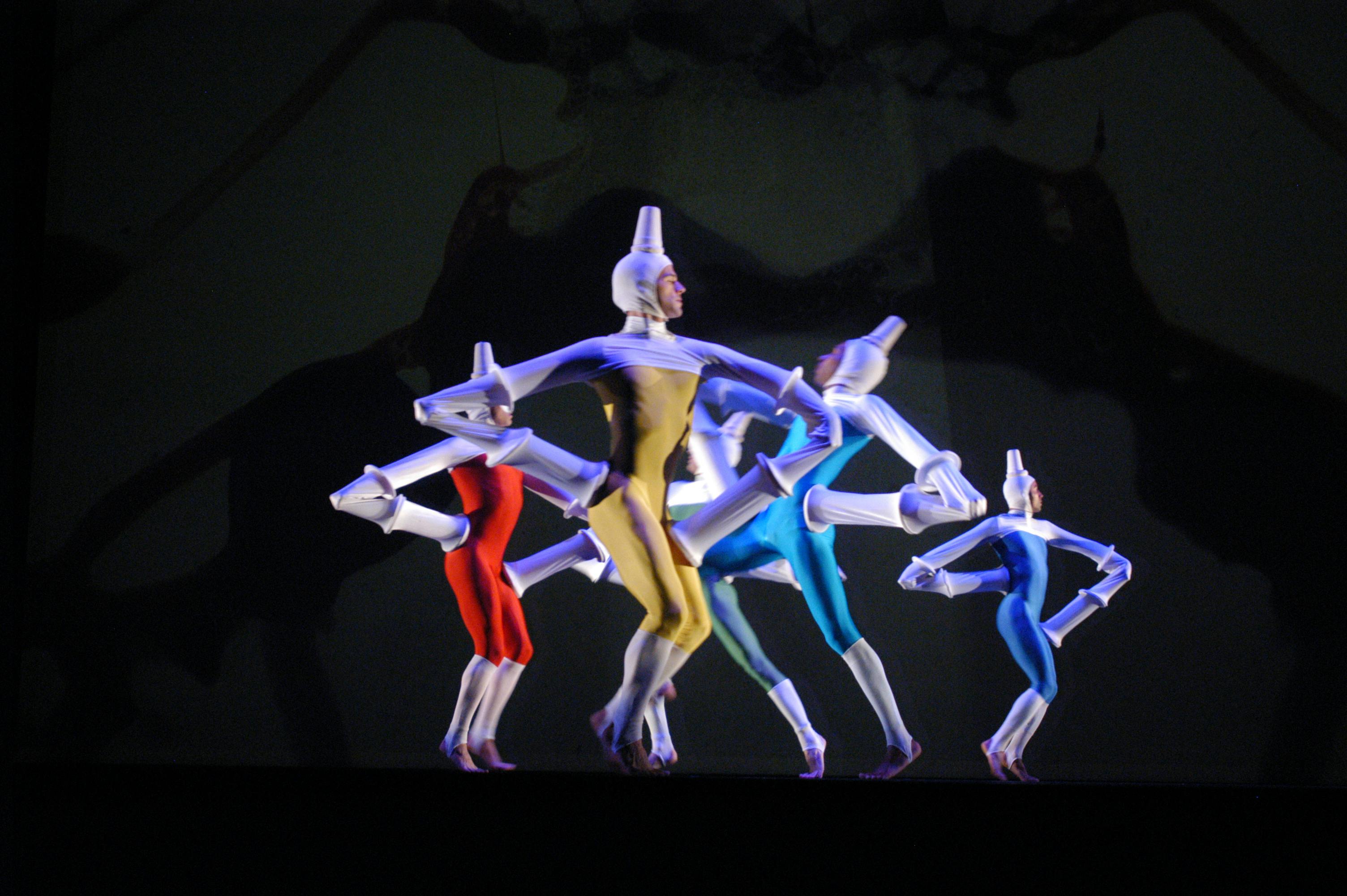 Four artists are on stage under black light. They wear tight-fitting suits and pointed headdresses.